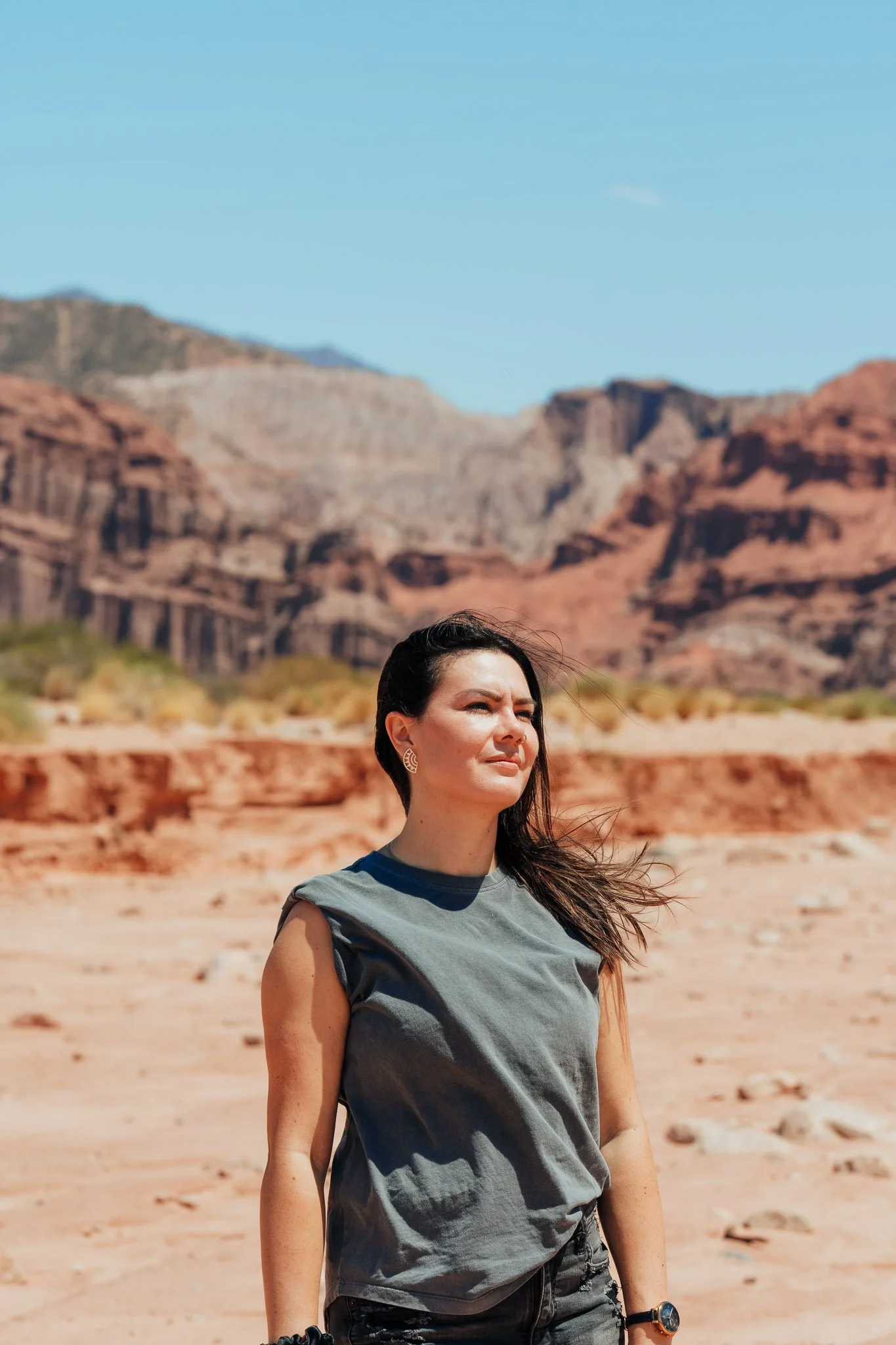 A woman with dark hair and earrings, wearing a gray sleeveless shirt and black shorts, stands outdoors in a desert with red rock formations and mountains in the background, under a clear blue sky.