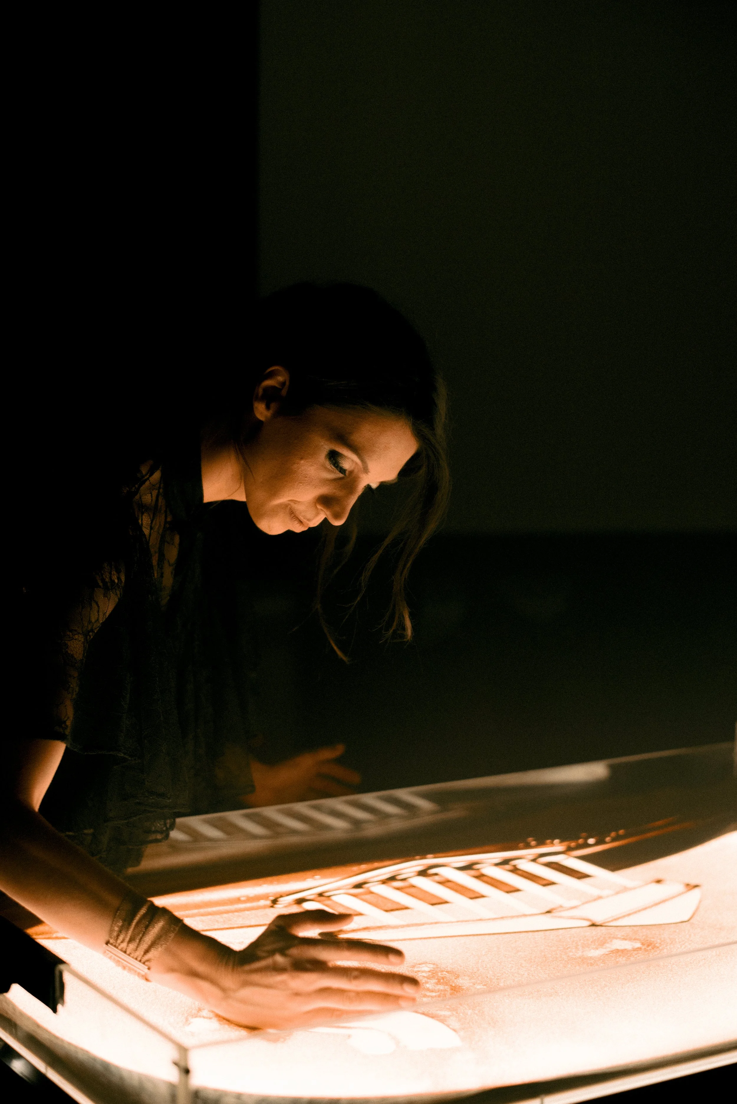 Nadia Pretto chinata sul piano luminoso mentre disegna nella sabbia, nel buio