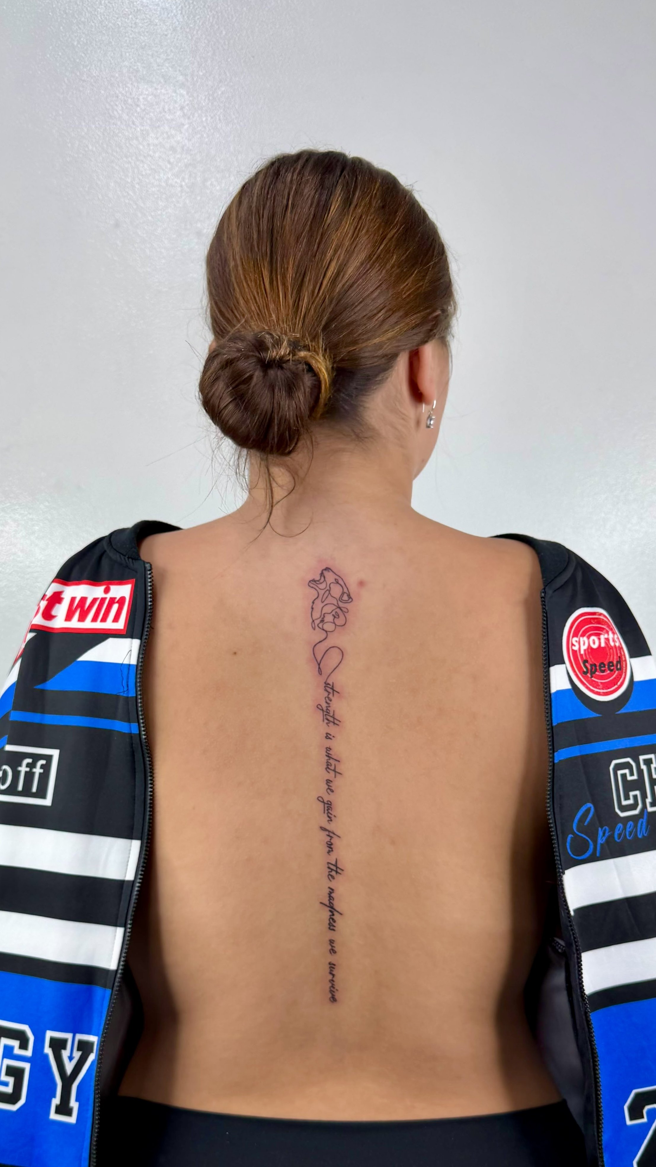 Back of a woman with a tattoo on her spine, wearing a sports jacket with various logos and patches.