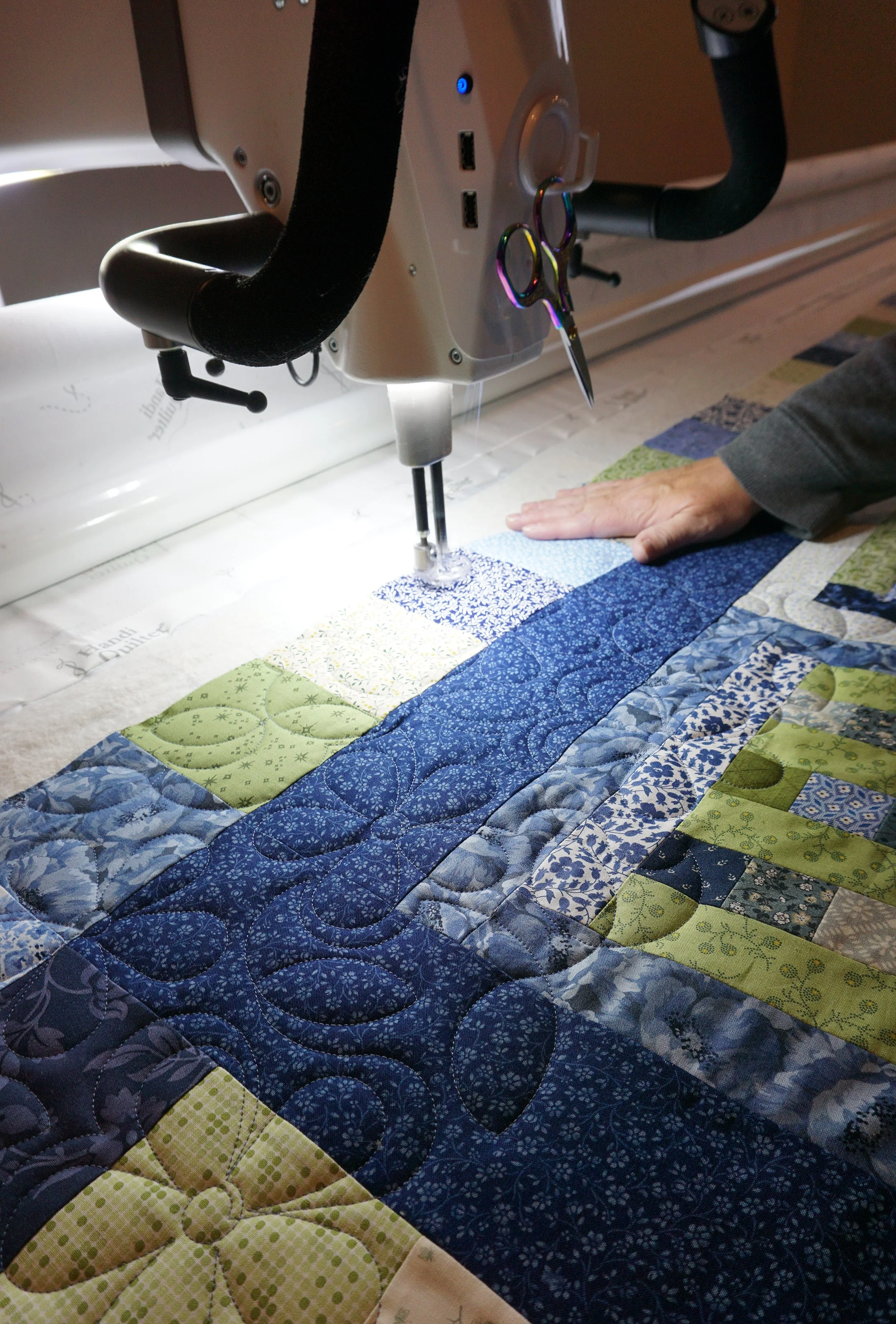 Close-up of a sewing machine quilting a colorful patchwork quilt with a person's hand guiding the fabric.
