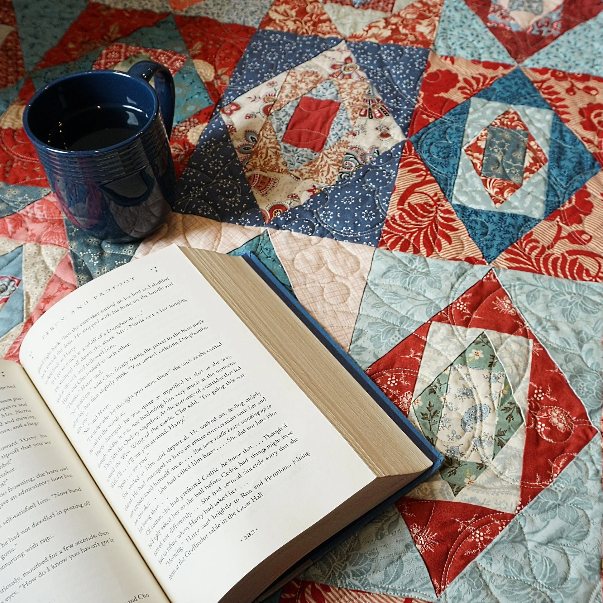 Open book with text on pages placed on a patchwork quilt featuring red, blue, and beige fabric patterns. A dark blue mug filled with a dark beverage is partially visible next to the book.