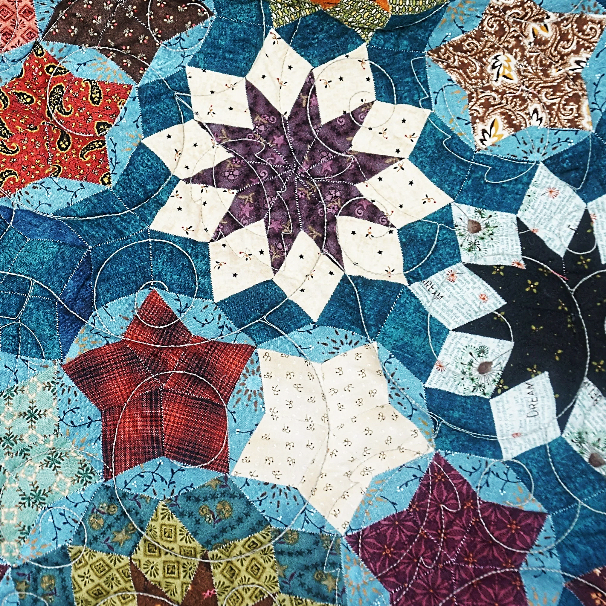 Close-up view of a colorful quilt with various patterned fabric patches sewn together, featuring a mix of floral, geometric, and paisley designs in colors like red, blue, purple, white, and brown.