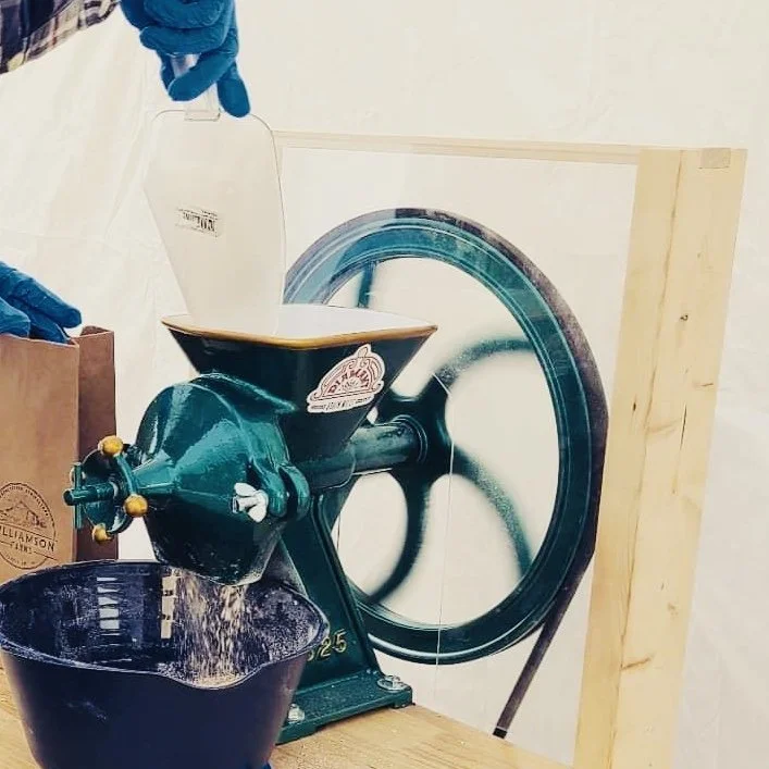 A person wearing blue gloves using a machine in a workspace, with a large paper or print of a wheel or gear in the background, and a brown paper bag nearby.