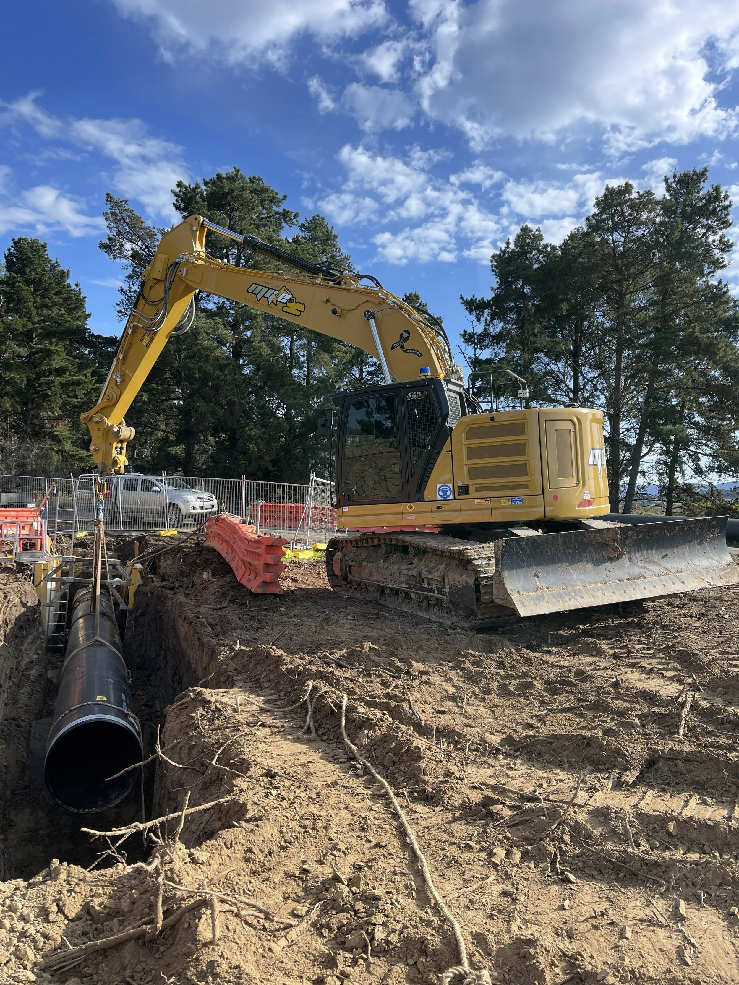 Marc Gray Earthmoving working on underground piping. The pipeline runs through the dirt in the foreground, with construction barriers and fencing nearby. 