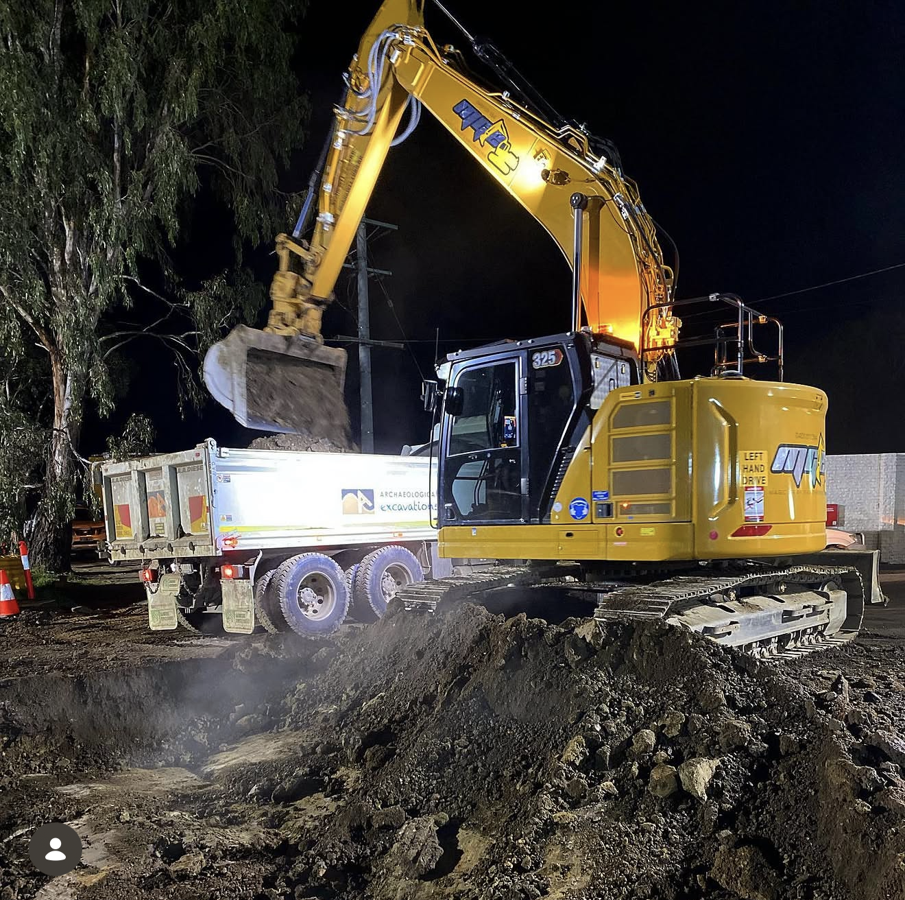 An excavator with a yellow arm is digging soil at night. It is unloading dirt into a truck labeled 'archaeological excavation' next to a tree and orange traffic cones.