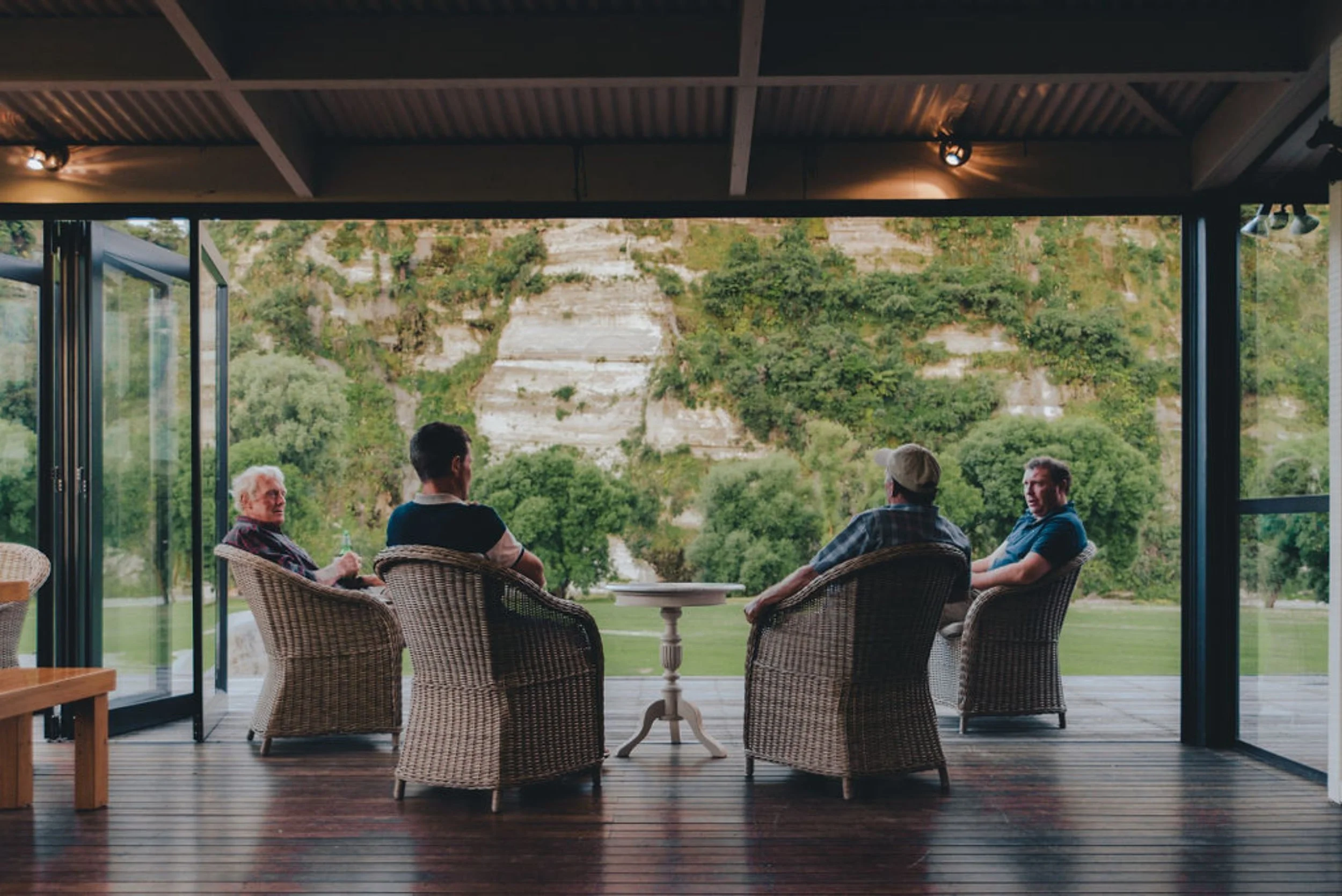 A group gathering at accommodation on a working farm, seated around a table on the porch or patio or Riverside Lodge, looking outside at a lush green landscape with trees and white cliffs.