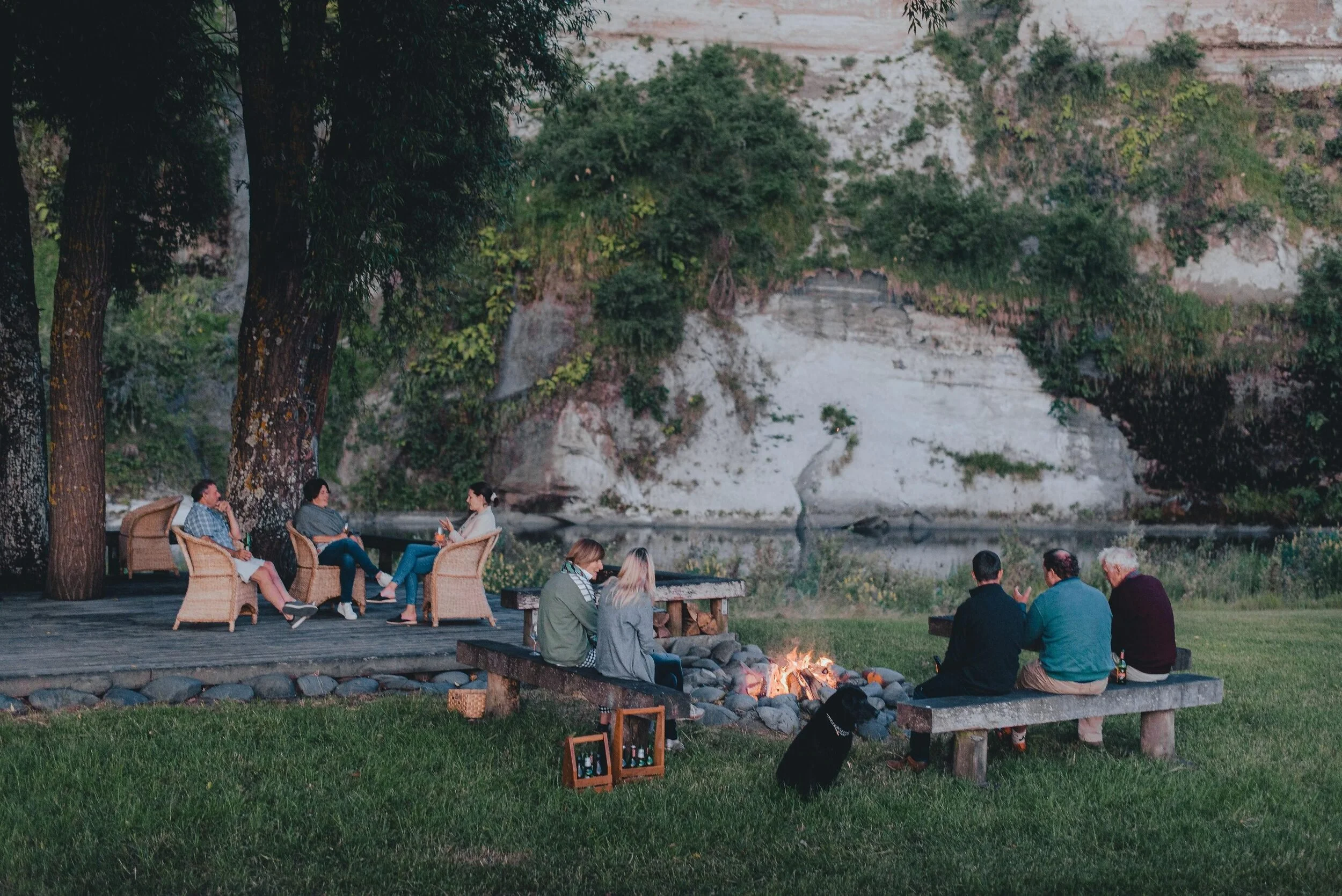 People sitting around a campfire on a grassy area, surrounded by trees, rocks and the Rangitikei River with the white papa cliffs in the background.