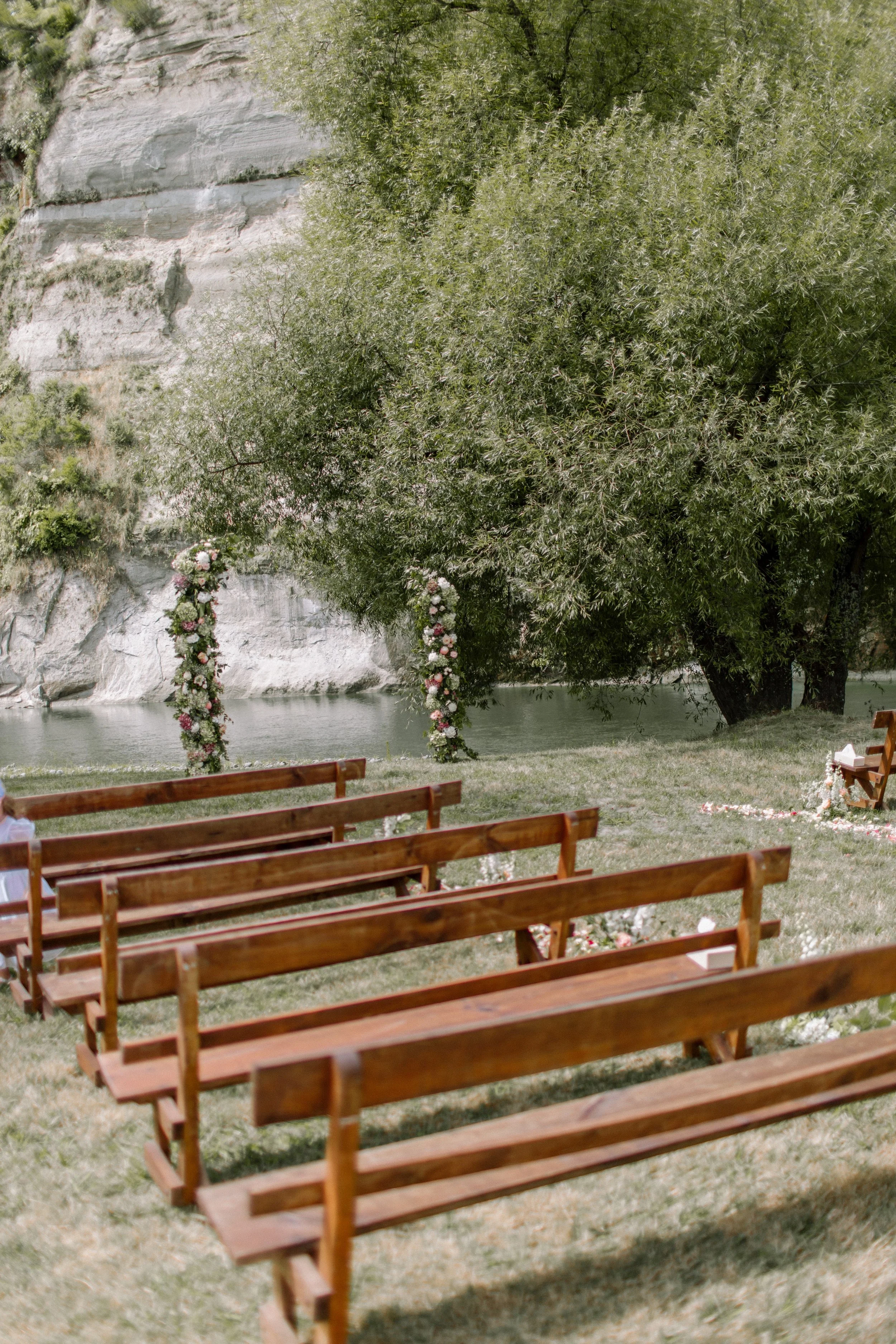 Outdoor wedding ceremony setup with wooden benches facing Rangitikei River, decorated with floral arrangements, under a large leafy tree near Manawatu.