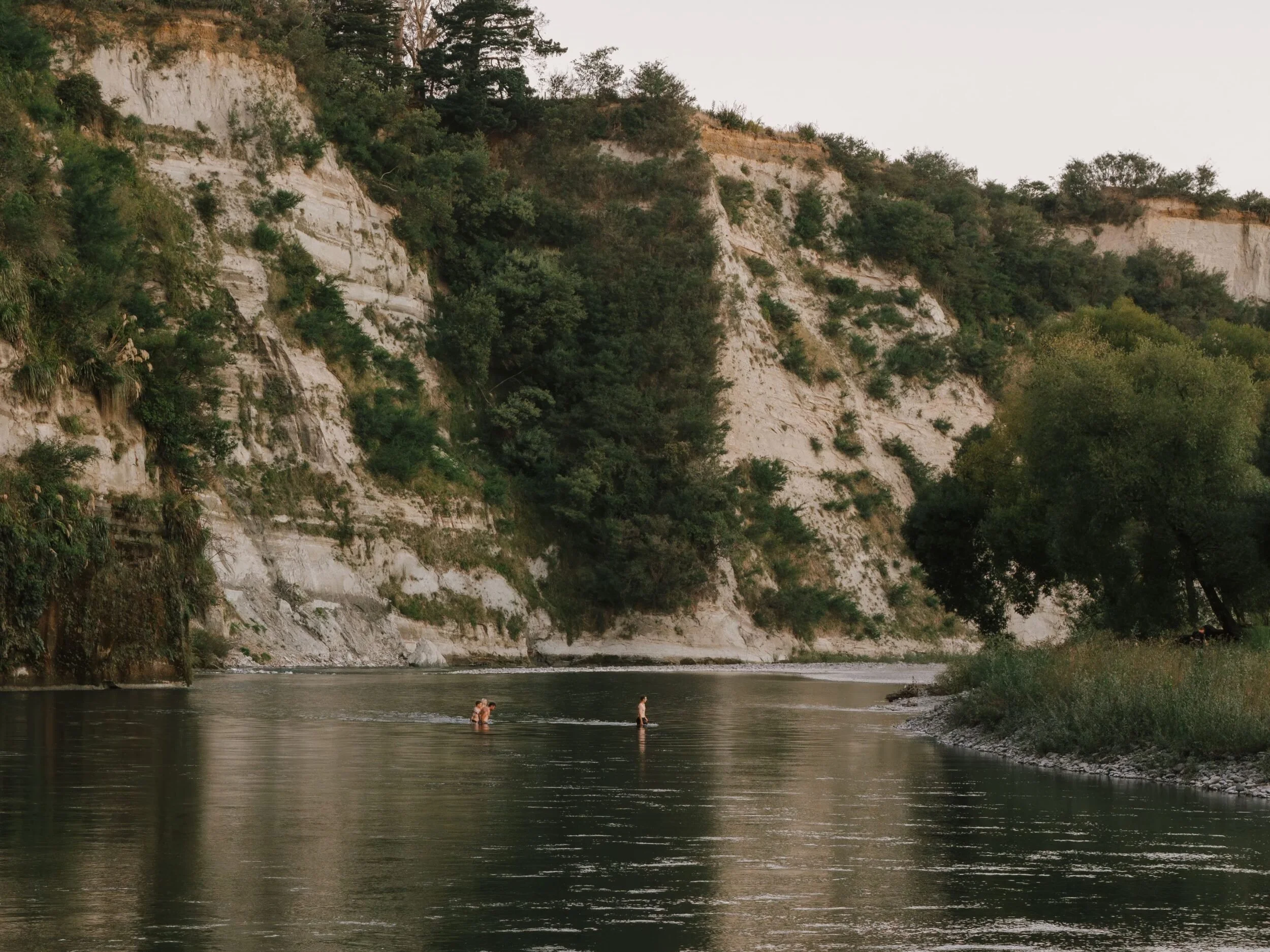 People enjoying a private swimming experience in Rangitikei River with cliffs and trees in the background during dusk.
