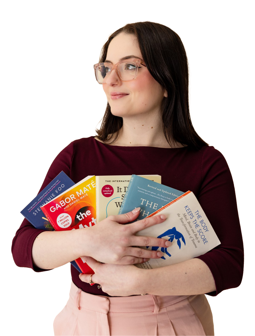 A young woman with glasses, wearing a burgundy top and pink pants, is holding several books and looking to her left.