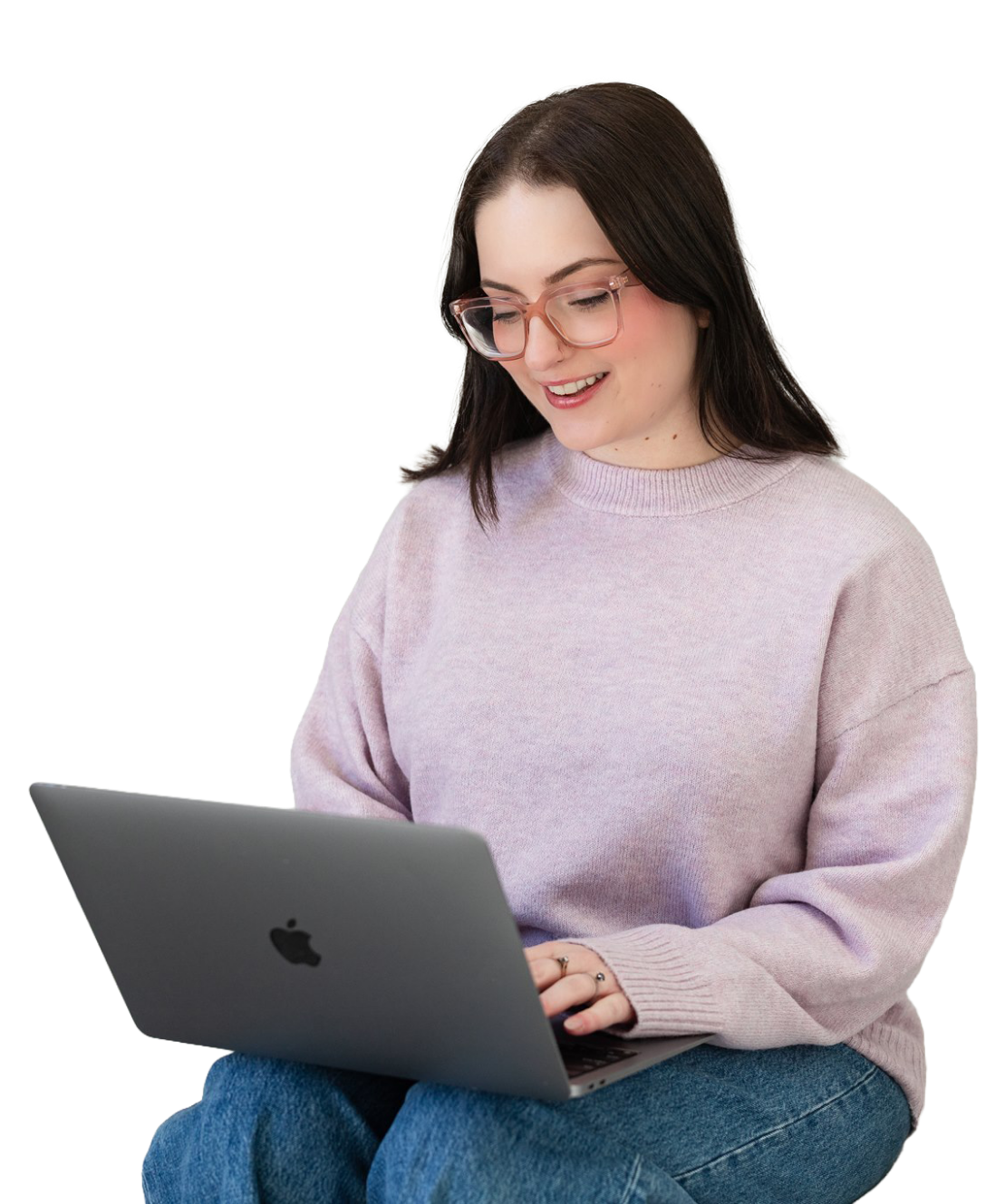 A woman with long dark hair, wearing glasses, a light pink sweater, and blue jeans, sitting and using a silver Apple MacBook laptop.
