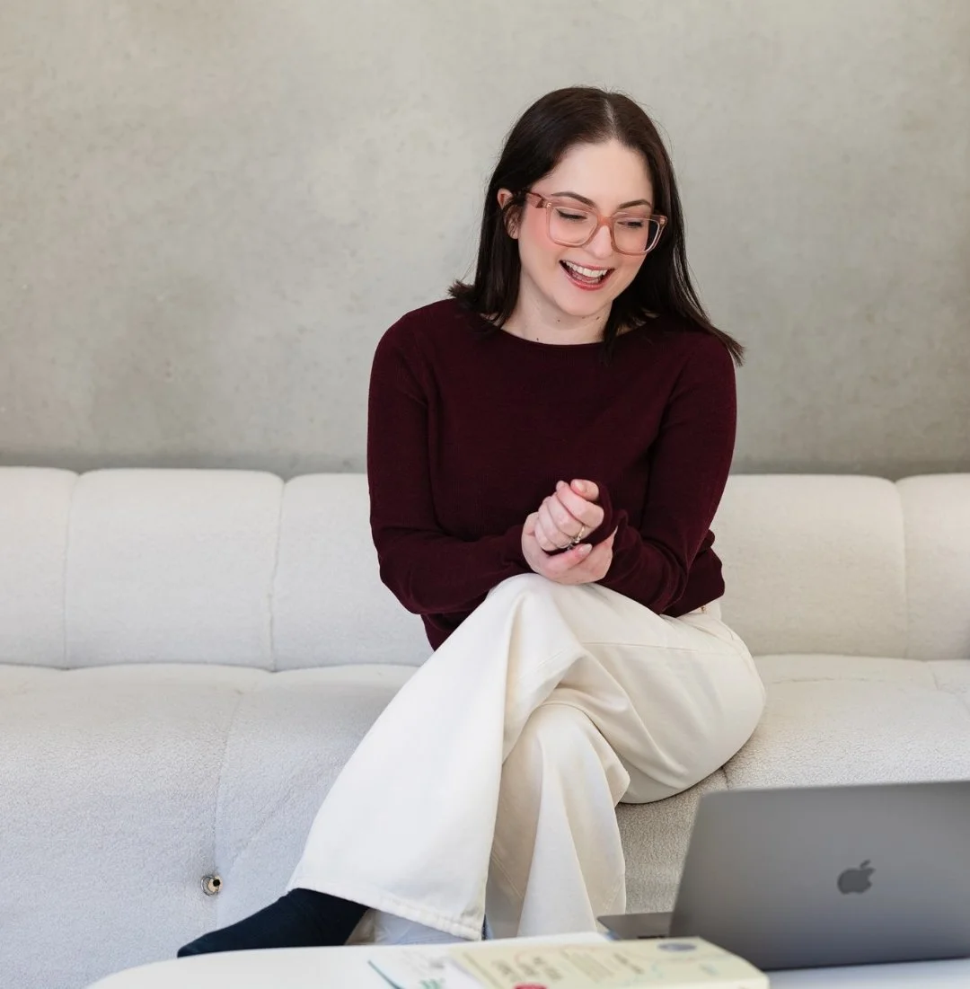 A woman with dark hair, glasses, and a burgundy sweater sitting on a white couch, smiling and looking down at her hands, with a laptop and some papers on a table in front of her.