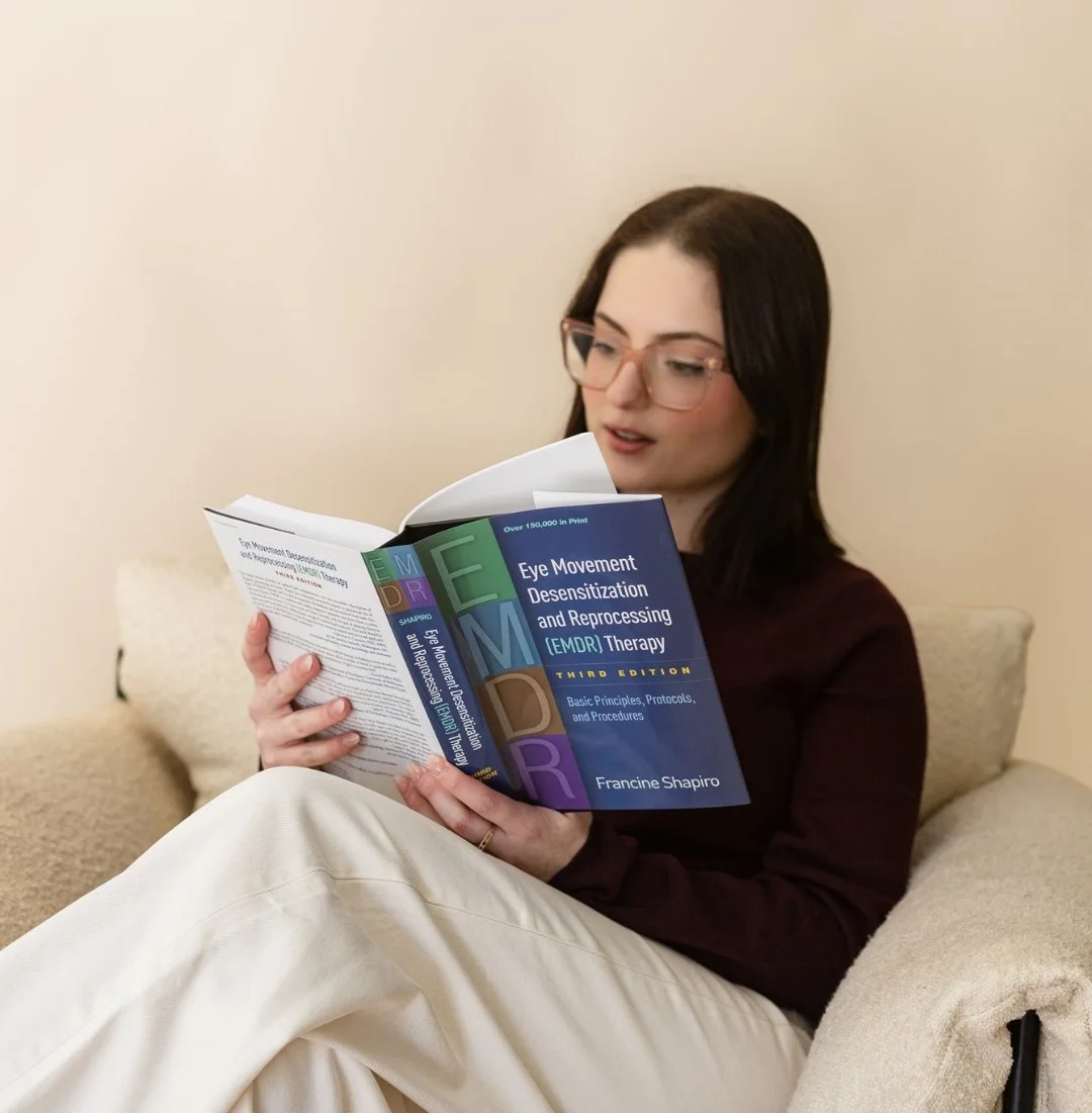 Woman with glasses sitting on a beige couch reading a book about EMDR therapy.