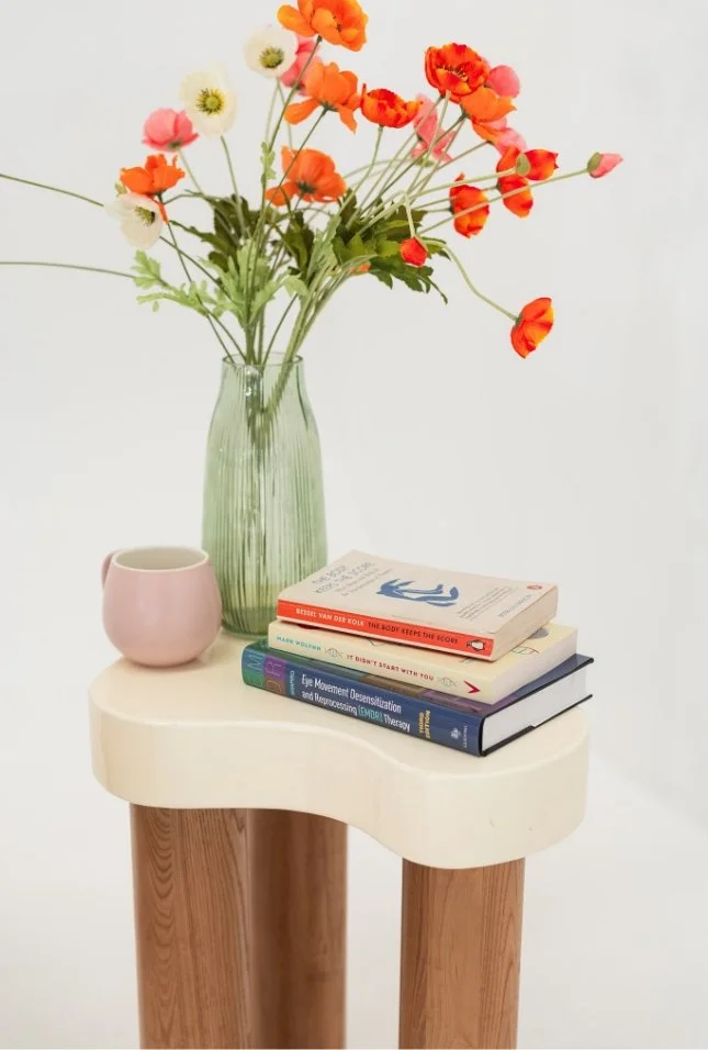 A small white side table with wooden legs holding a green vase with orange, white, and pink flowers, a pink mug, and a stack of five books against a plain white background.