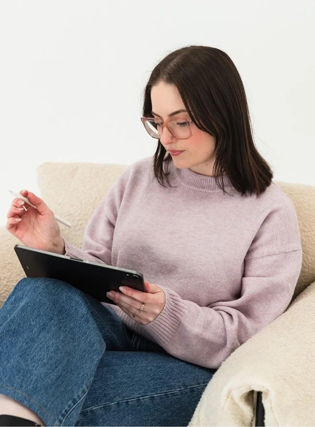 A woman with glasses wearing a pink sweater and jeans sitting on a beige couch, holding a pen and a drawing tablet, looking down at the tablet.