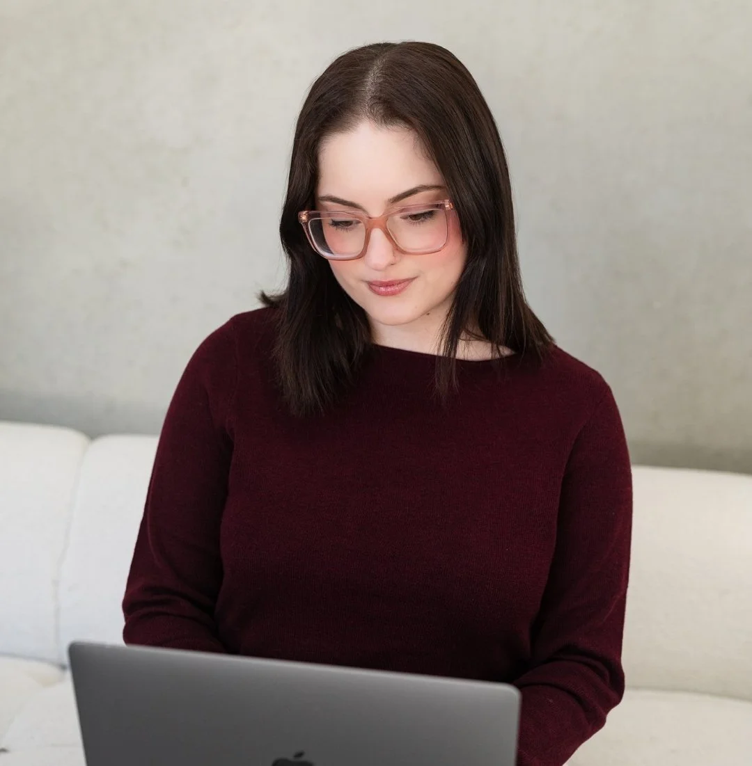 Young woman with dark hair and glasses using a laptop on a couch in a room with a plain wall.
