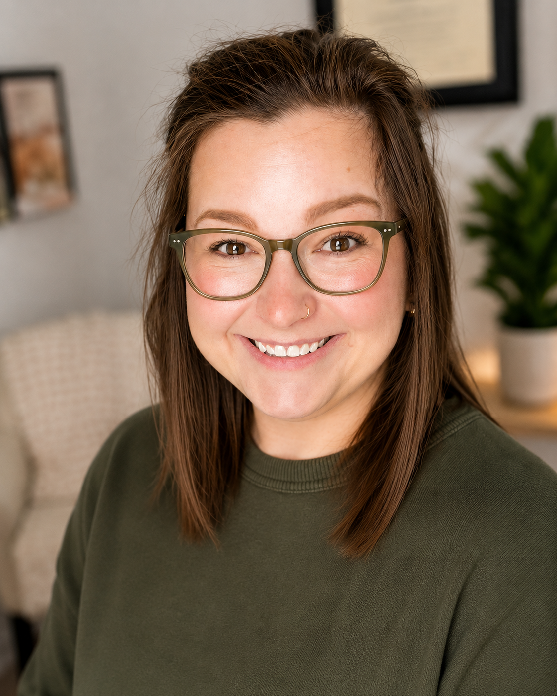 A woman with brown hair, wearing glasses and a nose ring, smiling at the camera in a cozy room with a beige chair and a potted plant in the background.
