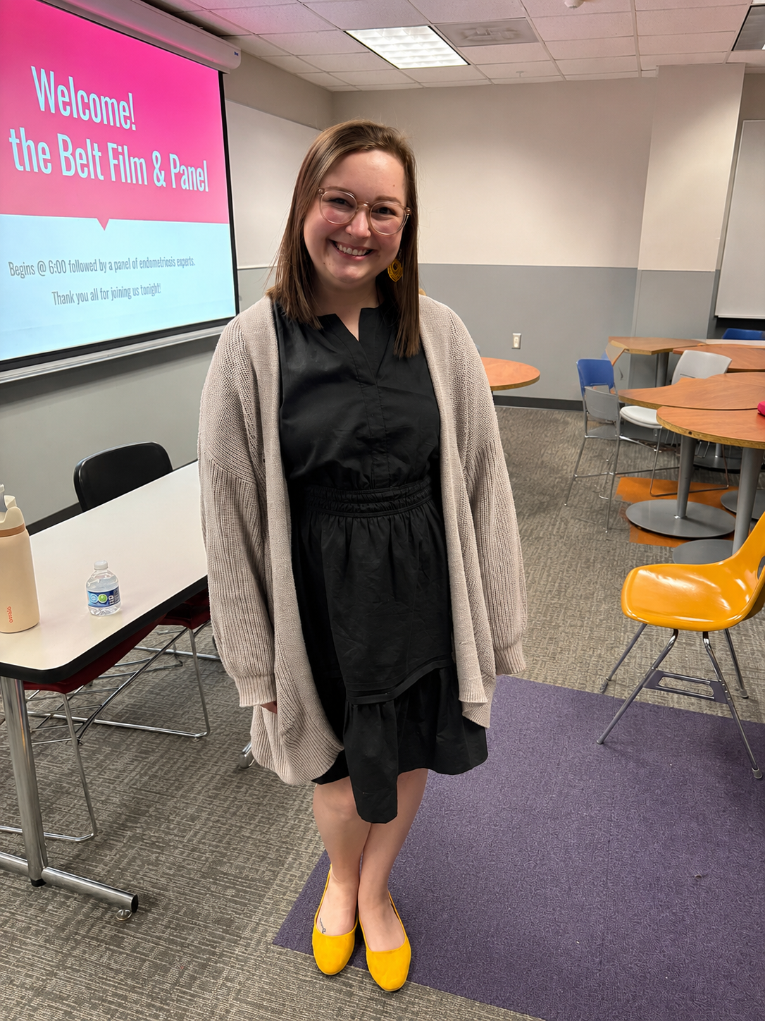 A smiling woman standing in a classroom or conference room with a presentation screen behind her. The screen displays a welcome message for the Belt Film & Panel event, with text indicating it begins at 6:00 pm and thanks attendees for joining. The room has tables and chairs, and the woman is wearing a black dress, a beige cardigan, and yellow shoes.
