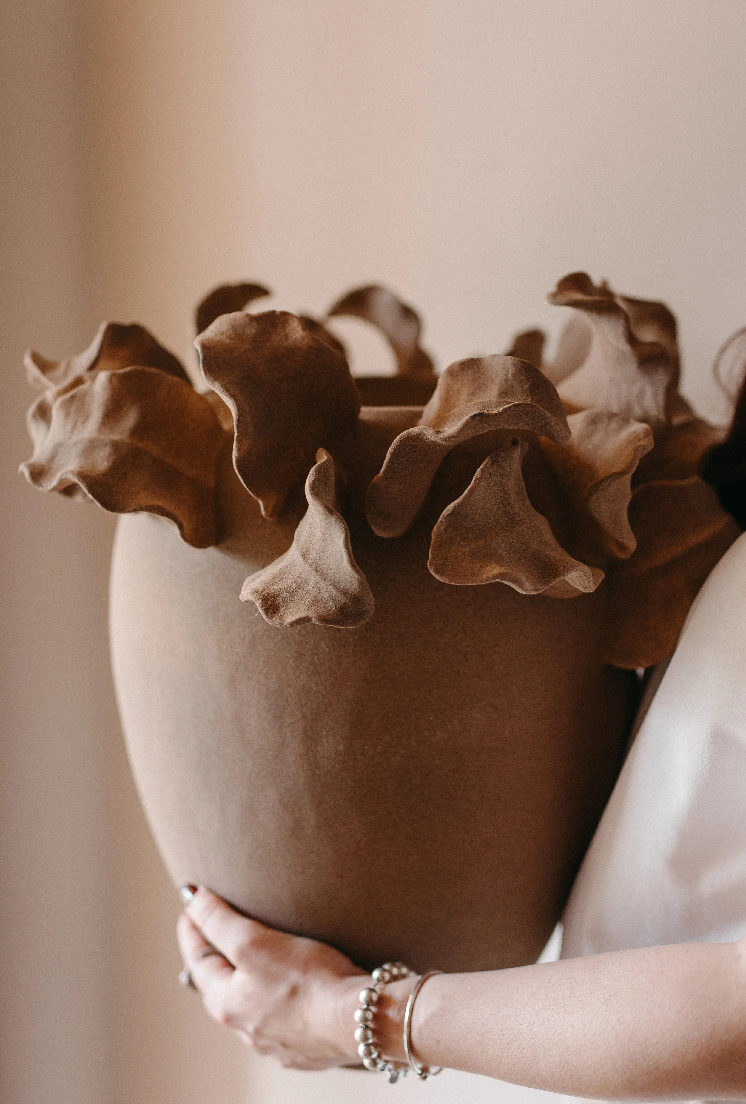 Person holding a brown ceramic vase with ruffled edges and dried leaves, wearing a peach-colored dress and silver jewelry.