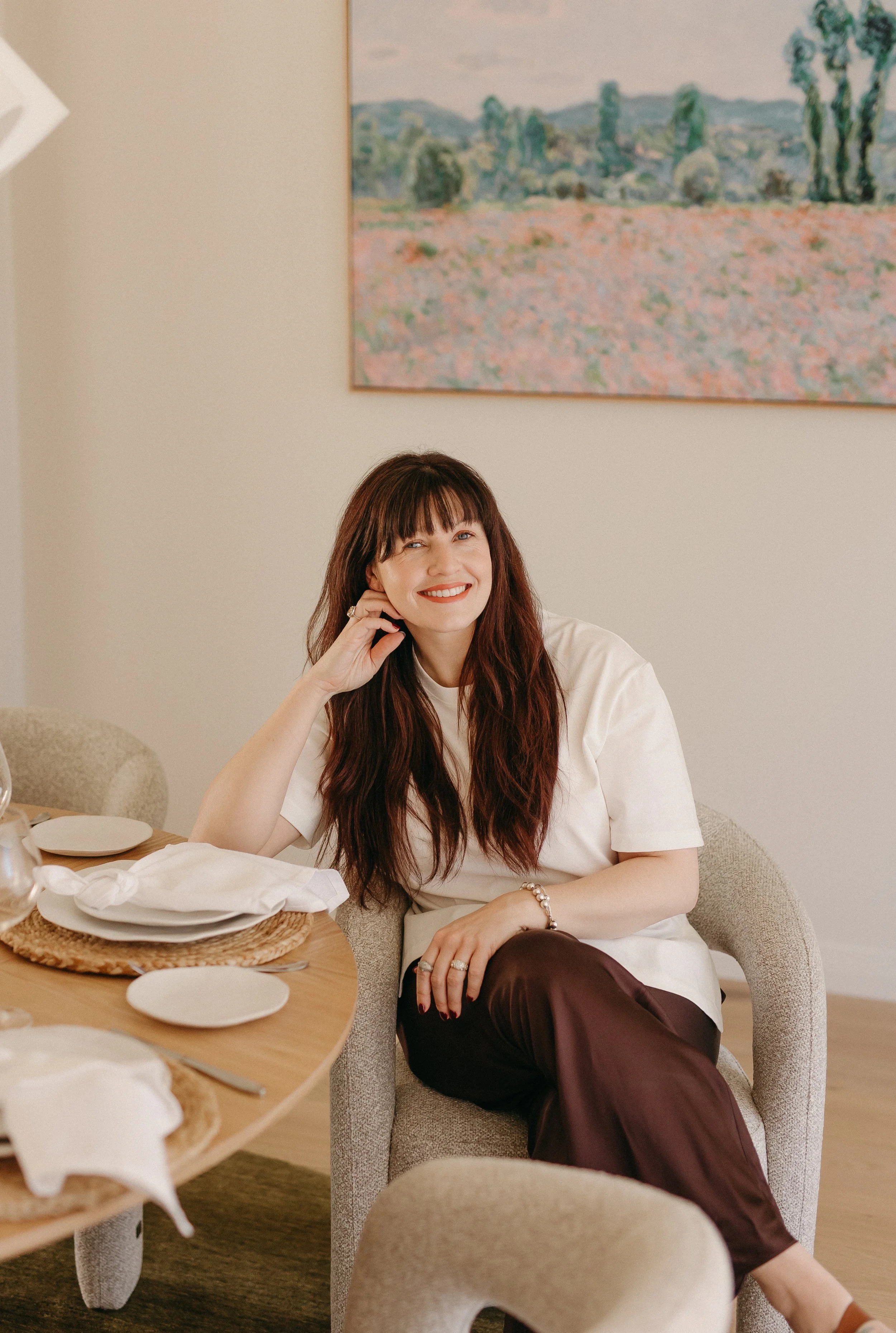 Kara, a woman with long brown hair and bangs sitting at a dining table, smiling, wearing a white top and dark brown pants. There are plates and utensils on the table, and a painting of a landscape is hanging on the wall behind her.