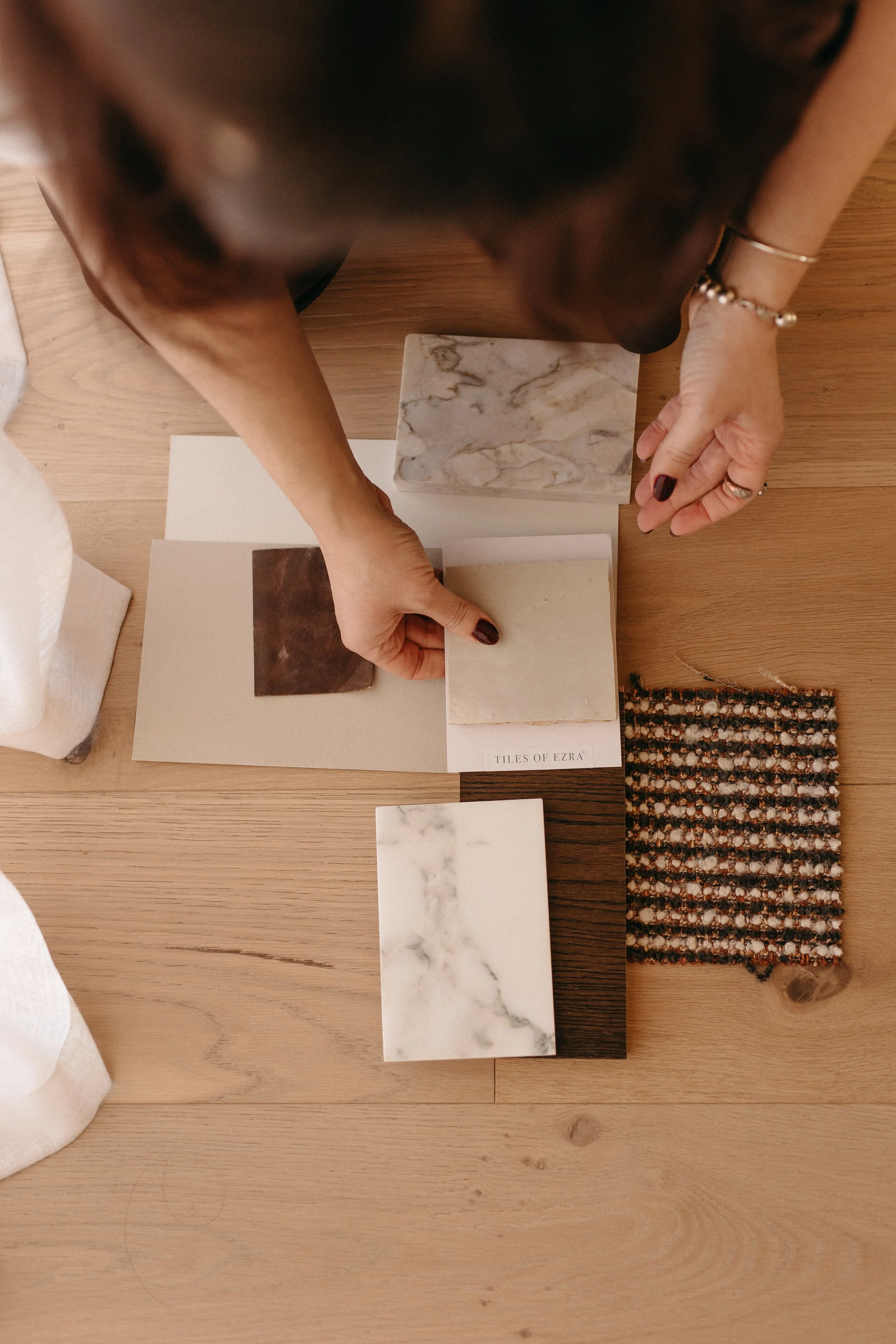 A person with dark painted nails and rings on their fingers is holding sample tiles with various marble and stone patterns. The tiles are spread out on a wooden table, with a small woven rug at the edge of the table. The person's face is not visible, and the focus is on the hands and the tiles.