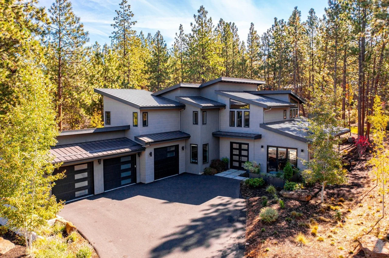 Modern house in a forested area with a driveway and landscaped yard, featuring multiple level roofs and large windows.