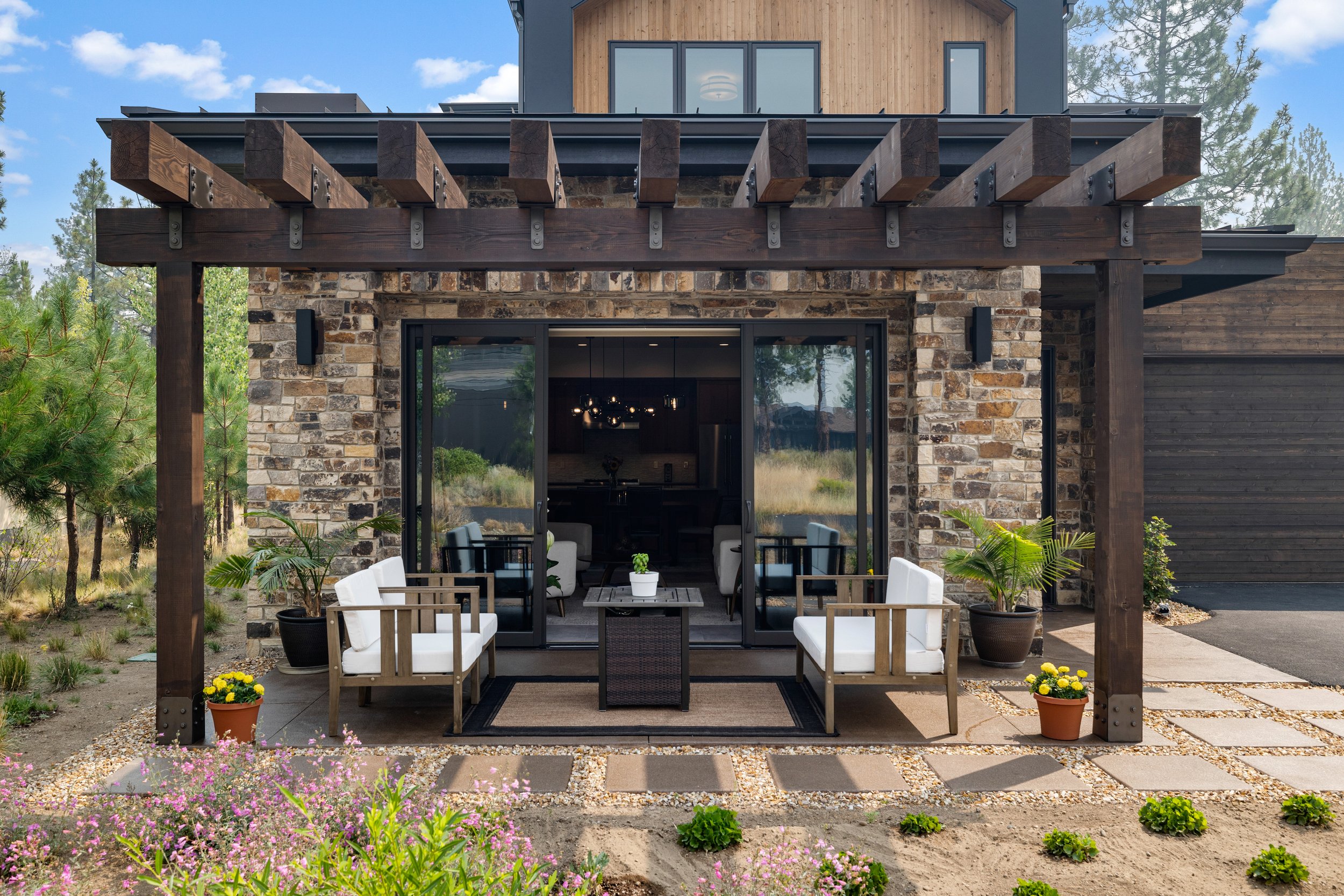 A modern house with a stone and wood exterior, large glass sliding doors, and a wooden pergola above an outdoor patio. The patio has white chairs, a small table, potted plants, and garden beds with flowers and greenery.