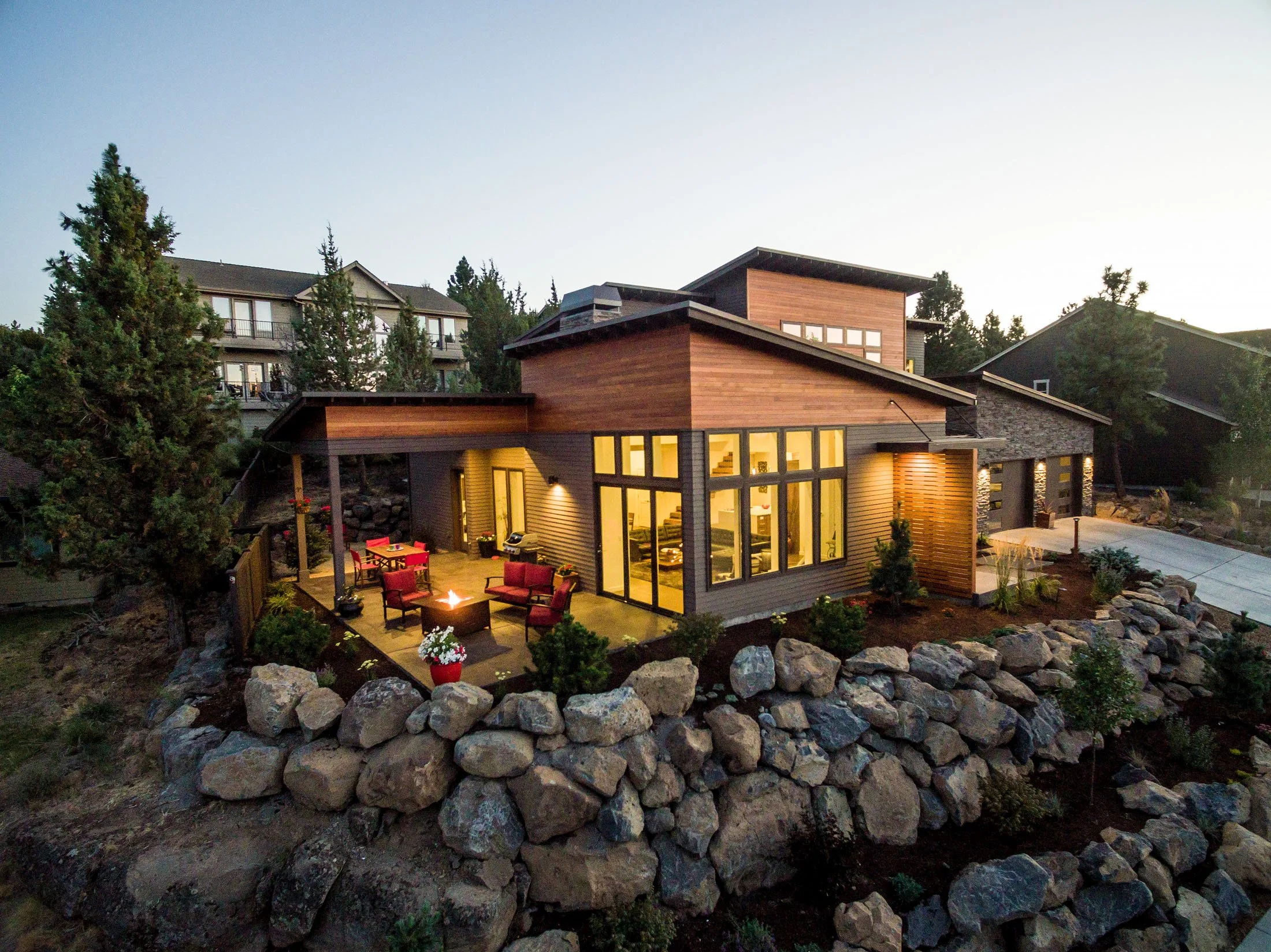Modern house with large windows and outdoor patio illuminated inside, surrounded by rocky landscape and trees at sunset.