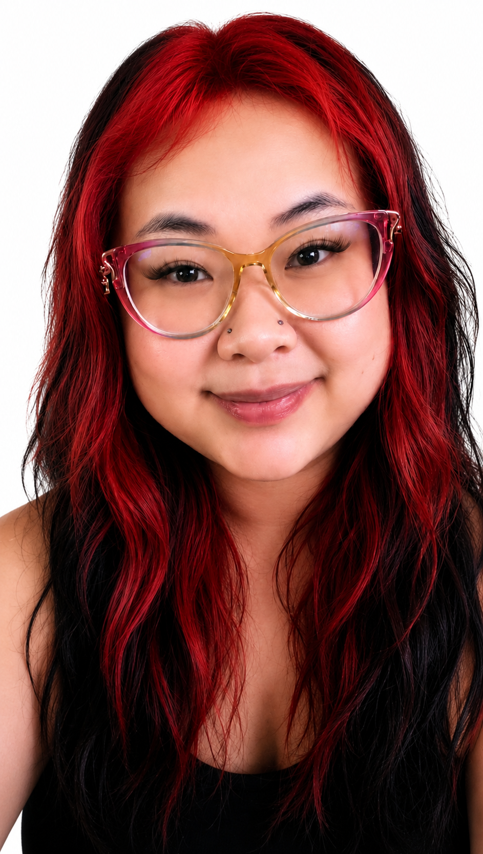Close-up portrait of a woman with red and black wavy hair, clear glasses with pink frames, and a nose piercing, smiling against a white background.