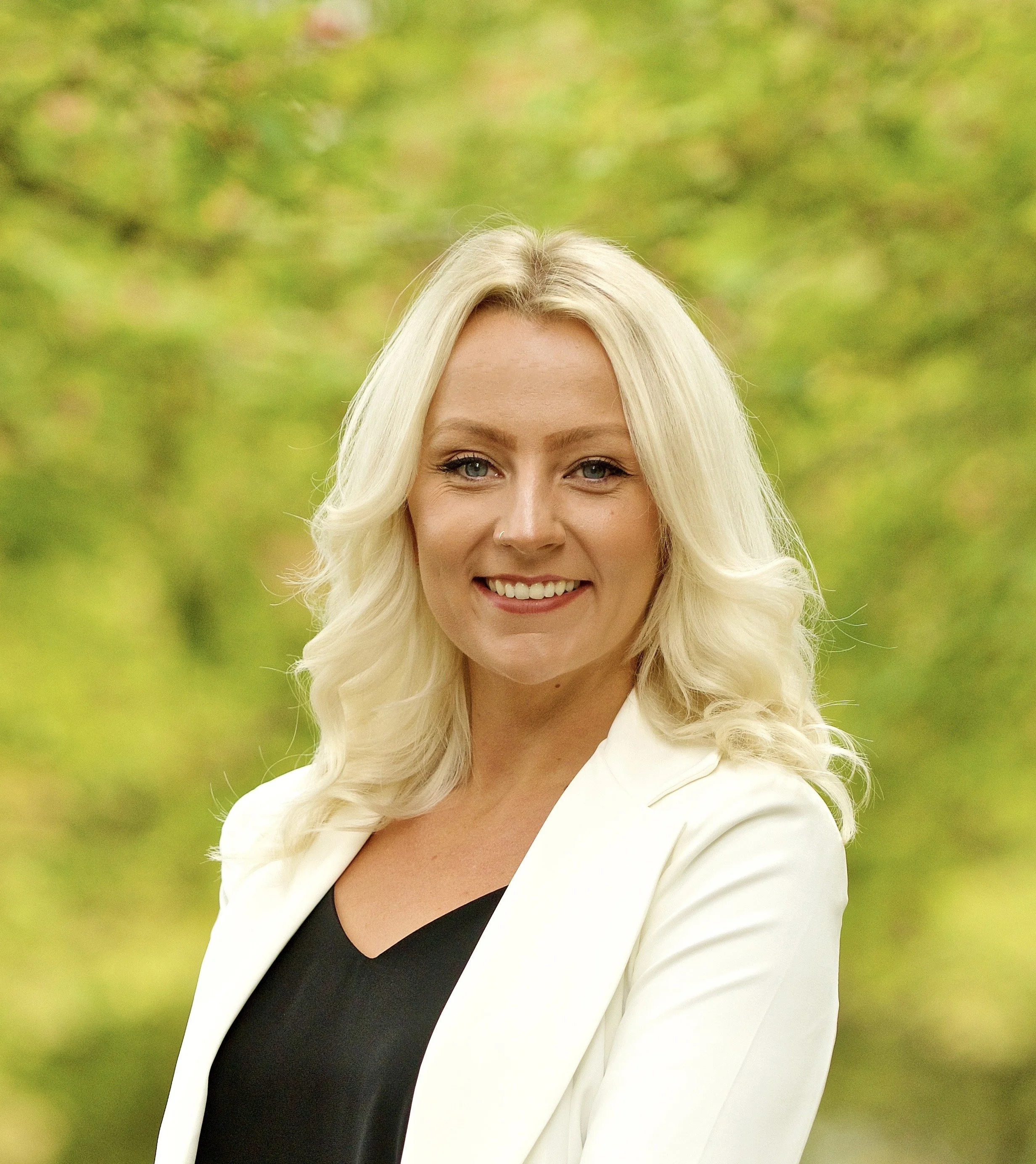 A woman with blonde hair, wearing a white blazer with black and a black top, smiling outdoors with a blurred green background.