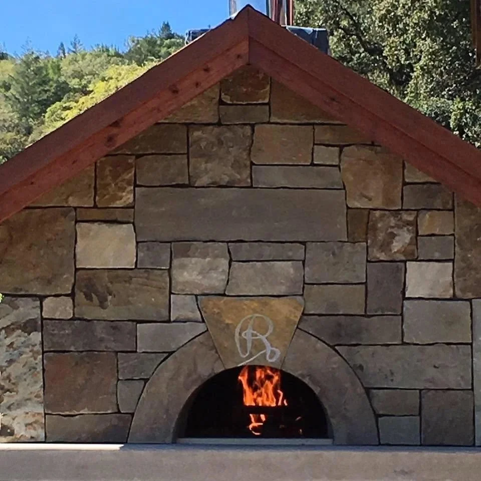 A stone fireplace with a wood mantel and a fire burning inside. The house has a gabled roof with red trim, surrounded by trees and a clear sky.
