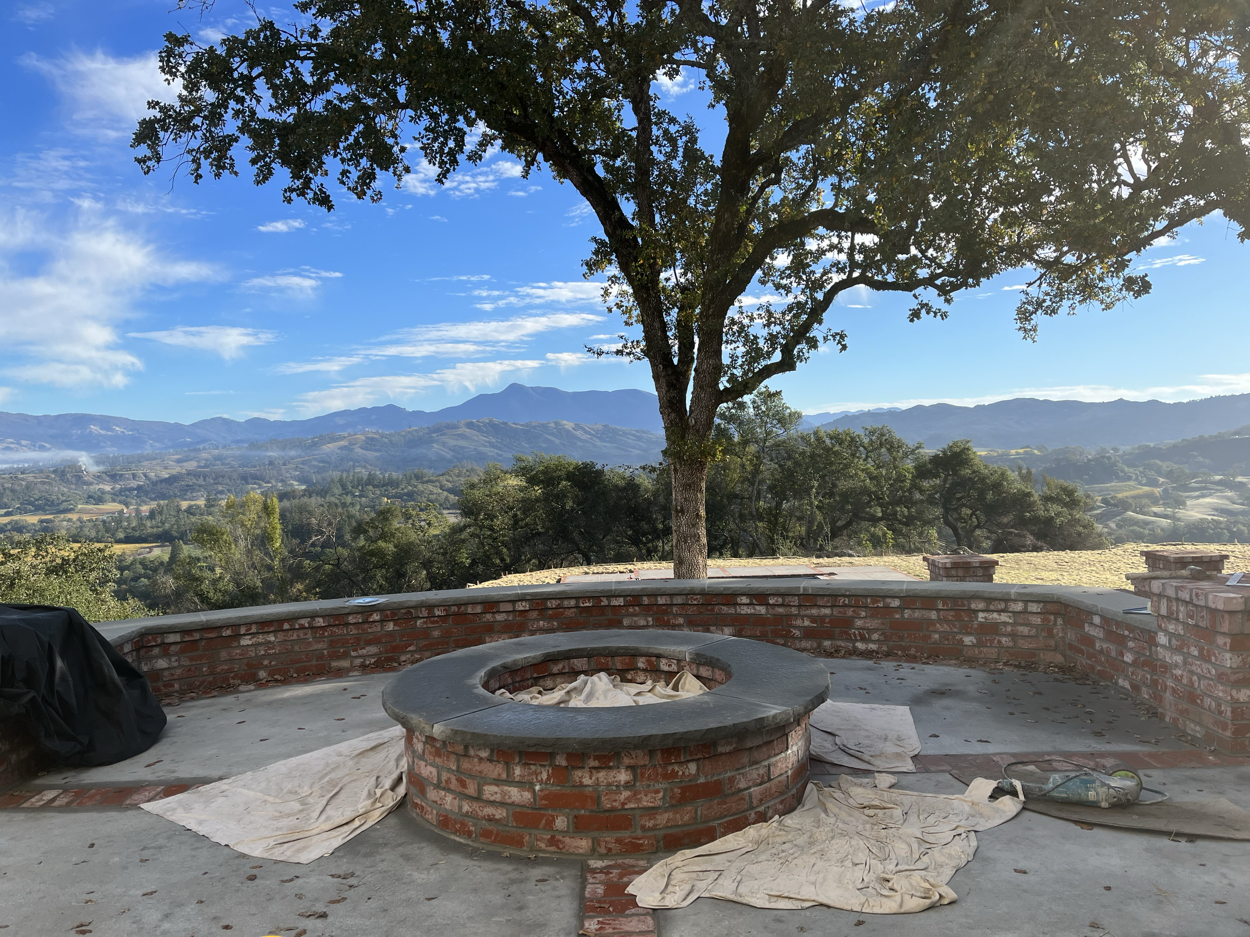 A scenic view of mountains in the background with a large tree in the foreground. In front of the tree, there is an outdoor brick structure that appears to be under construction or renovation, with some construction materials and fabric on the ground