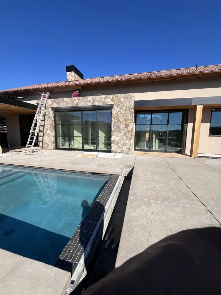 Construction workers installing large sliding glass doors on a house's back patio, with a swimming pool in the foreground and a ladder leaning against the house.
