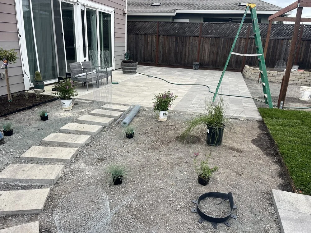 Backyard patio in progress with paving stones, a ladder, potted plants, and outdoor furniture.