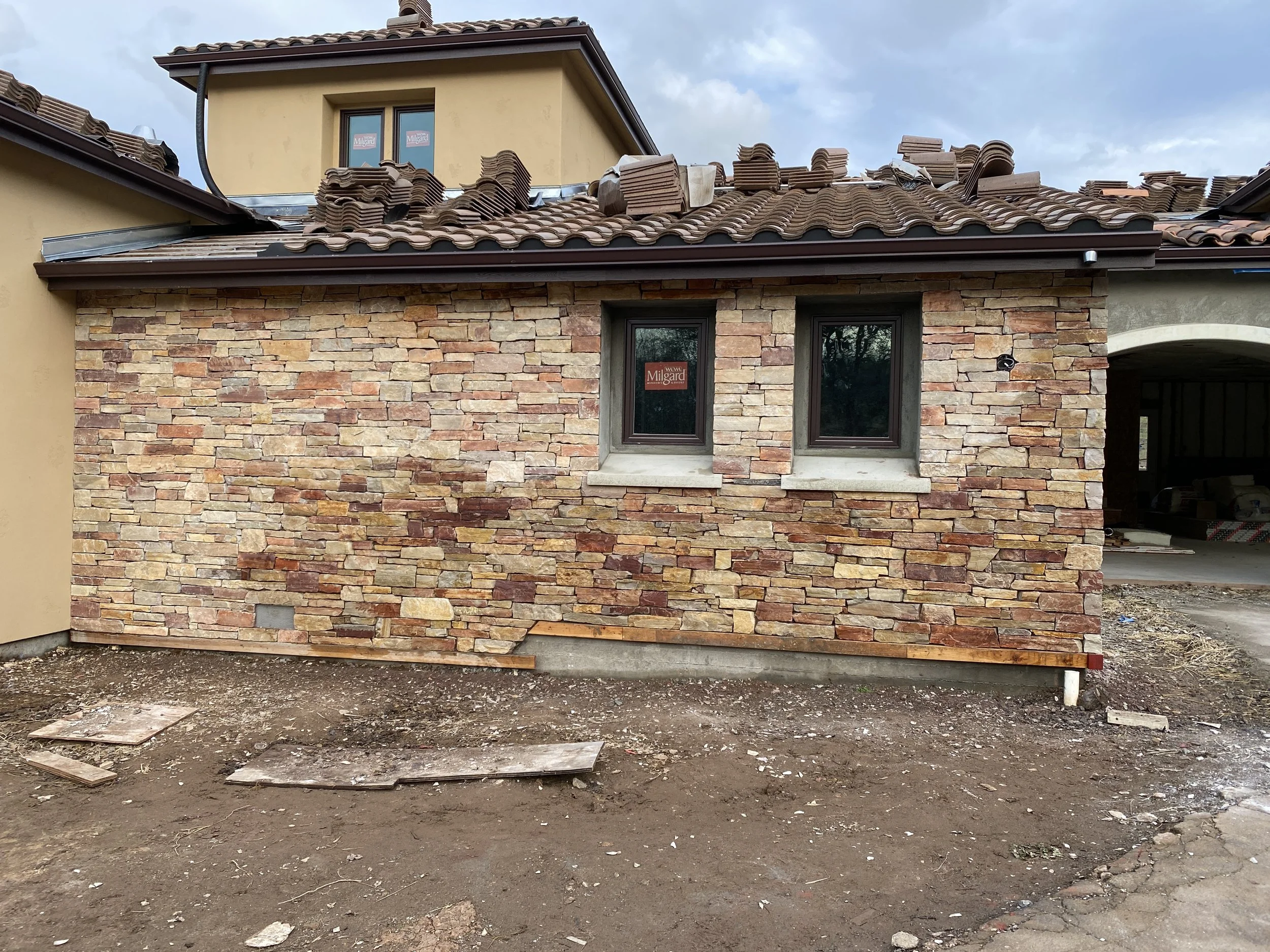 Partially constructed house with a brick exterior wall, a large window, and a roof with clay tiles, some of which are piled up on the roof, and construction materials on the ground.