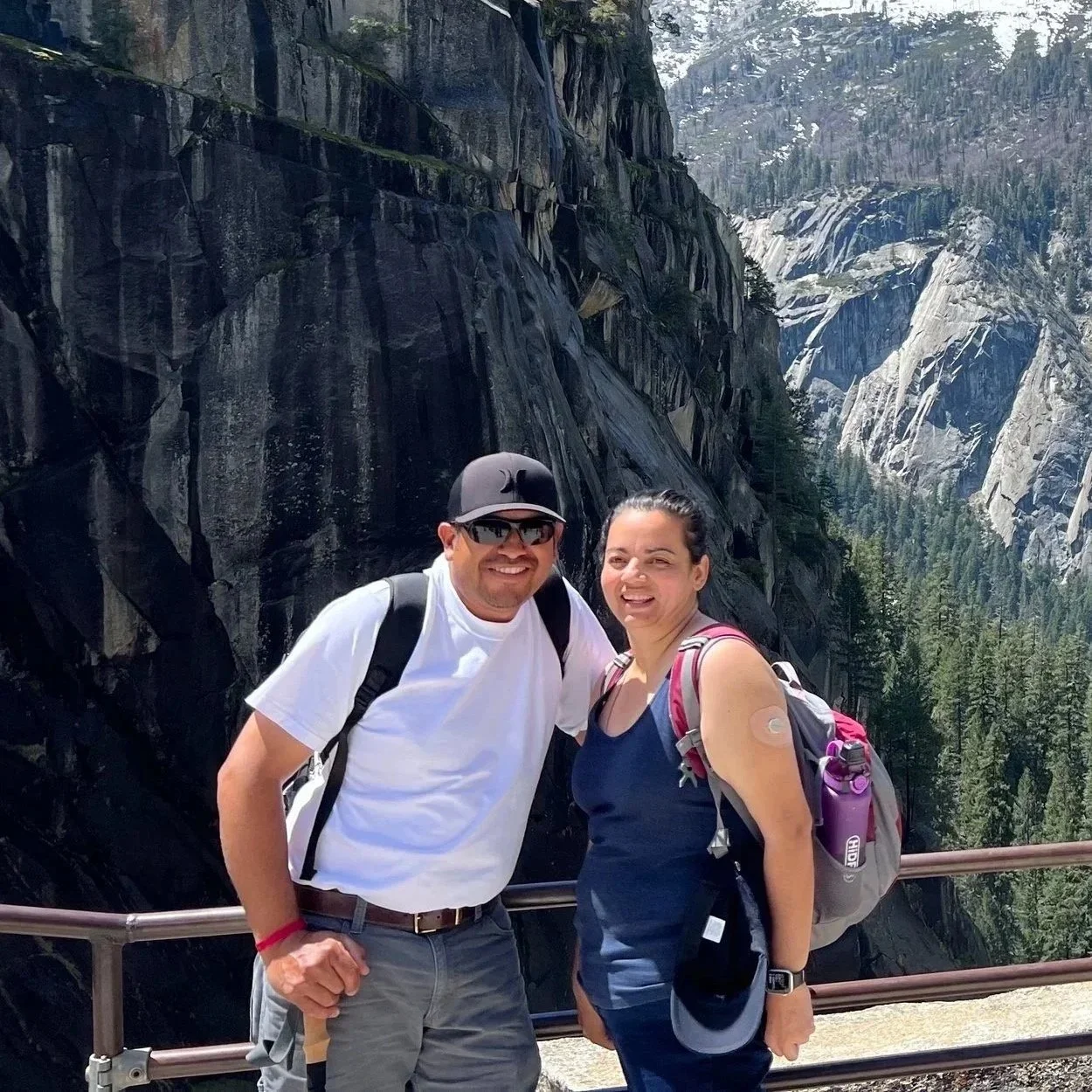 A smiling man and woman with backpacks posing on a mountain overlook with tall granite cliffs and forested valleys in the background.