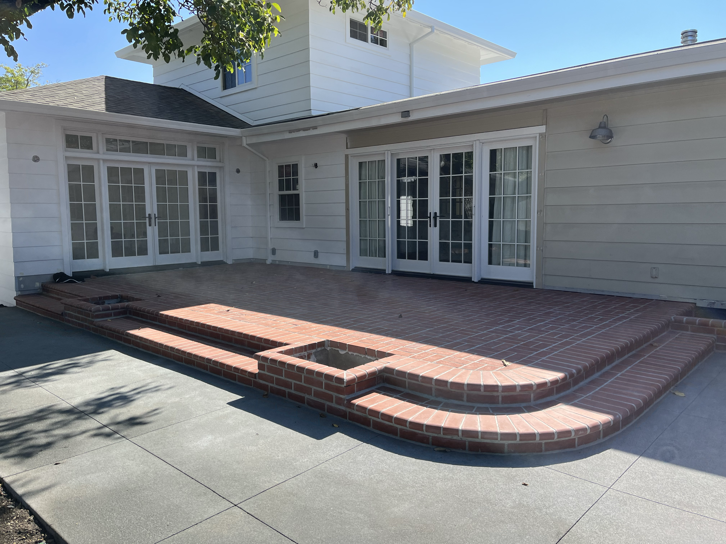 A backyard patio with brick steps and a concrete surface, connected to a white house with large glass sliding doors and several windows. There are trees overhead and a bright blue sky.