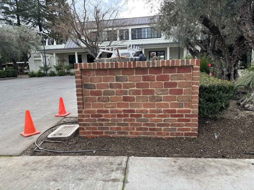 A brick wall with two orange construction cones placed on the sidewalk next to it, and a utility access panel nearby. In the background, there are two people working on equipment and a large residential building with trees and bushes surrounding the 