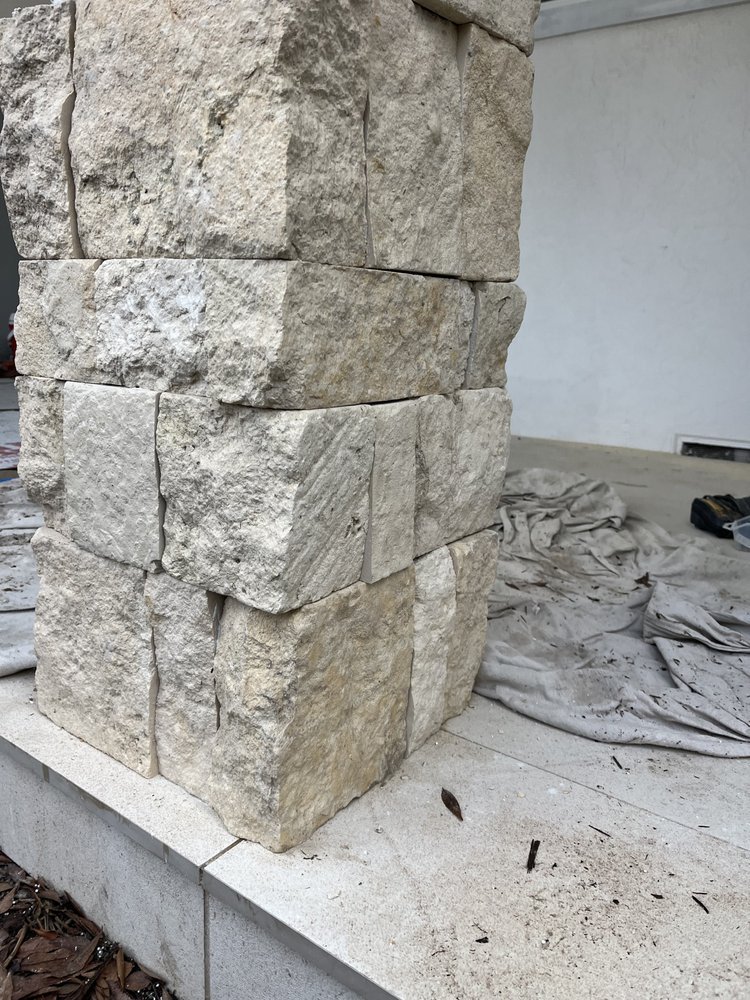 Close-up of a stone pillar with rectangular beige stones stacked on top of each other during construction, with construction materials and a tarp on the ground nearby.