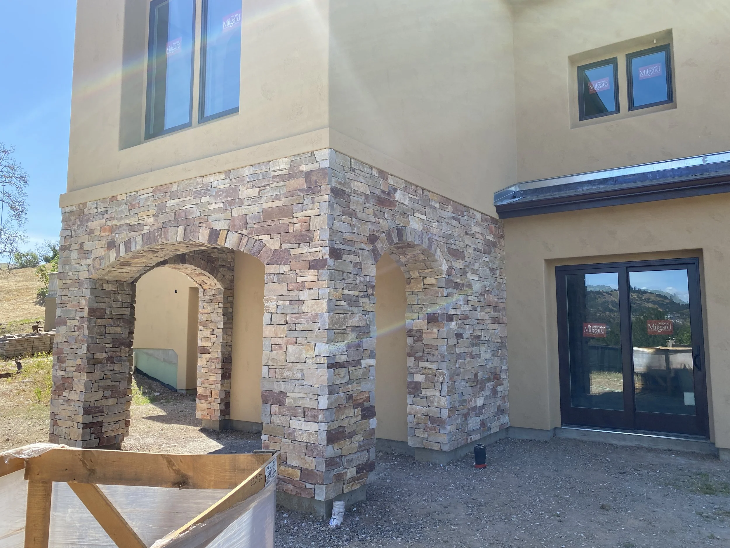 Construction site of a building with an upper wall painted beige and a lower section with stonework, including an arched opening and windows with stickers that say 'Milgard,' indicating ongoing window installation.