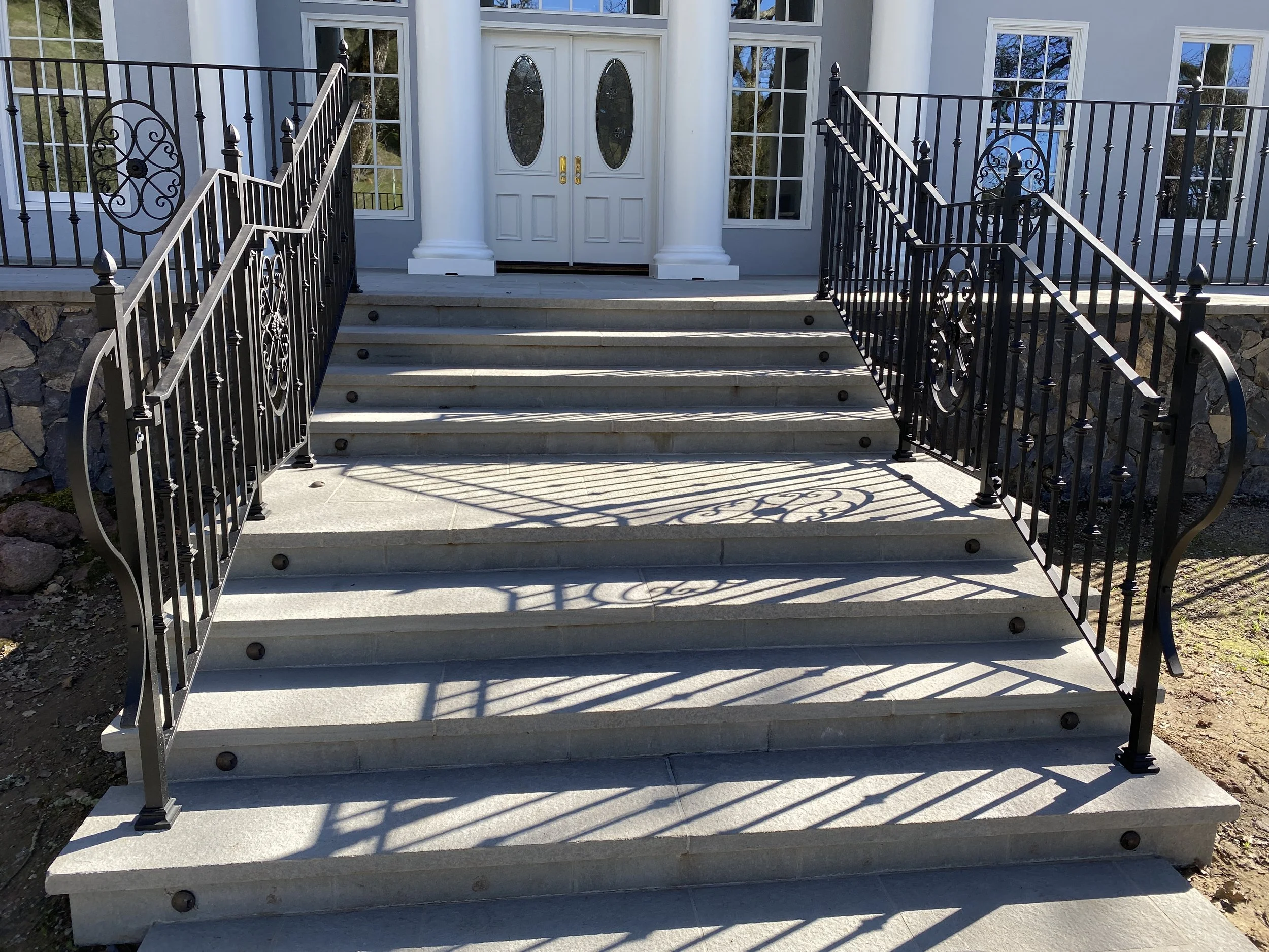 Front porch with gray steps, ornate black metal railings, and white double front doors with oval glass inserts, flanked by tall white columns.