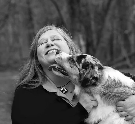 A woman smiling and holding a dog that is licking her face outdoors.