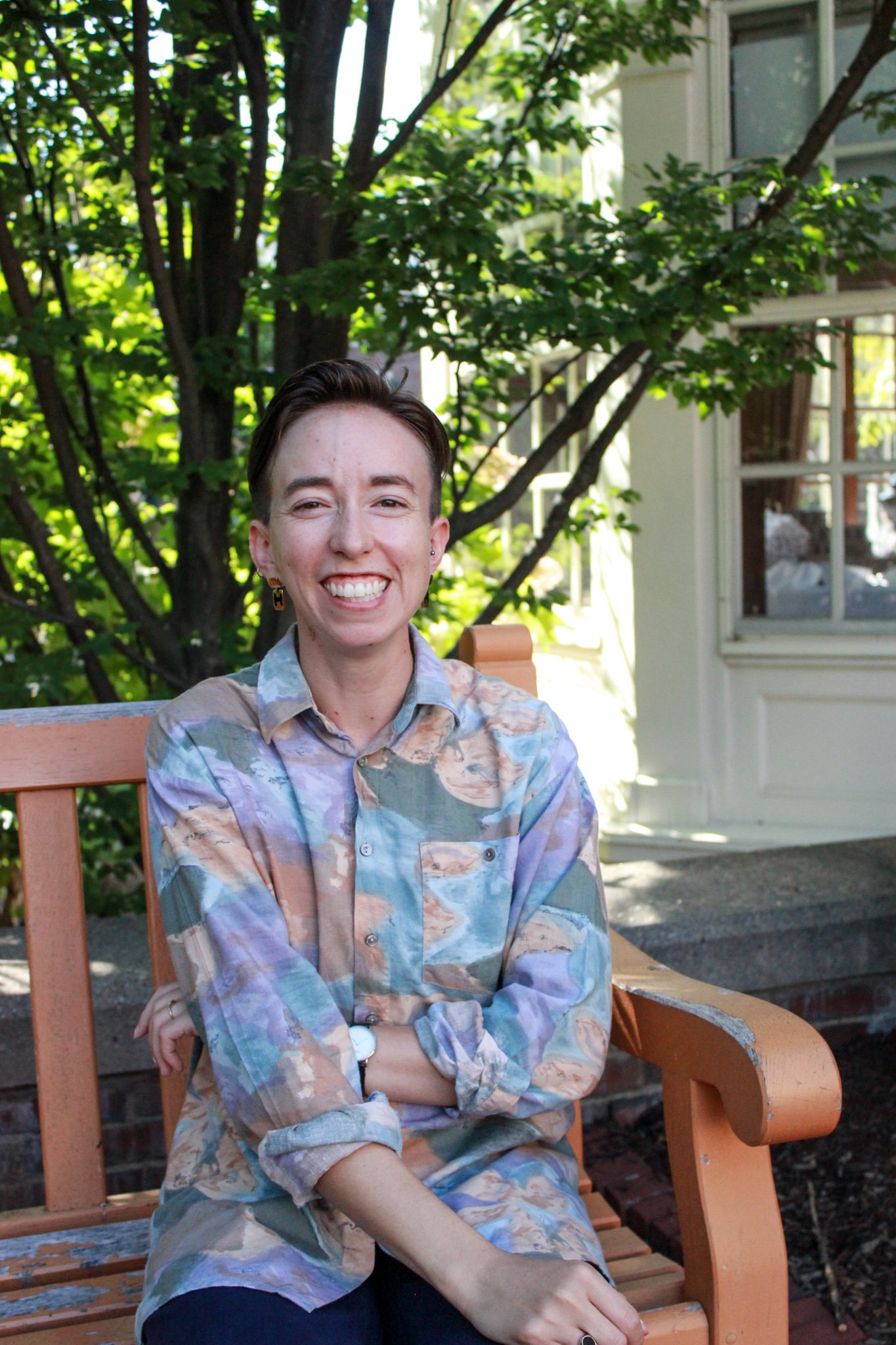 A smiling woman with short brown hair sits on an orange wooden bench outdoors, wearing a multicolored abstract patterned shirt, with a background of green leaves and part of a house window.