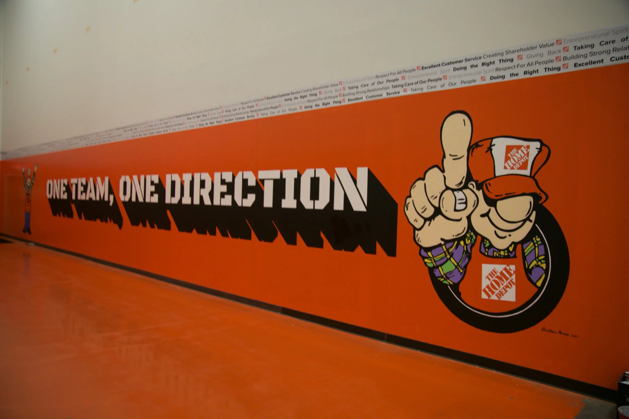 Orange wall featuring a cartoon figure of a hand pointing with two fingers, with the text 'ONE TEAM, ONE DIRECTION' and the logo of The Home Depot.