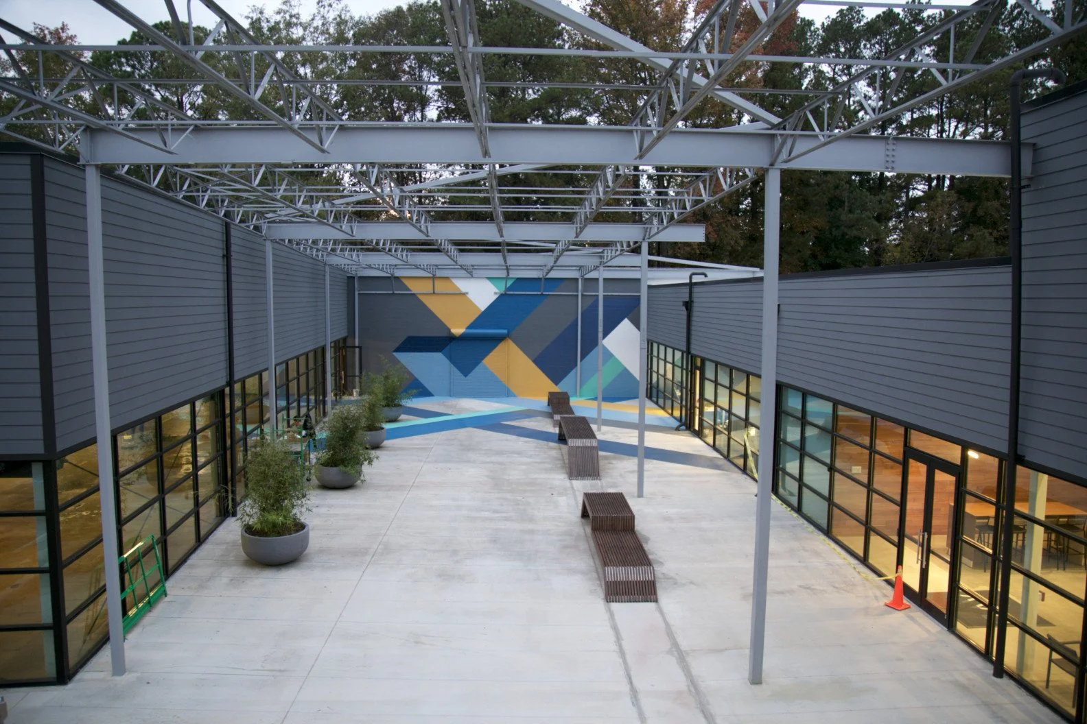 View of an outdoor terrace area with modern gray building walls on either side, a geometric mural on the far wall, potted plants, a sculpture, benches, and a steel framework overhead.