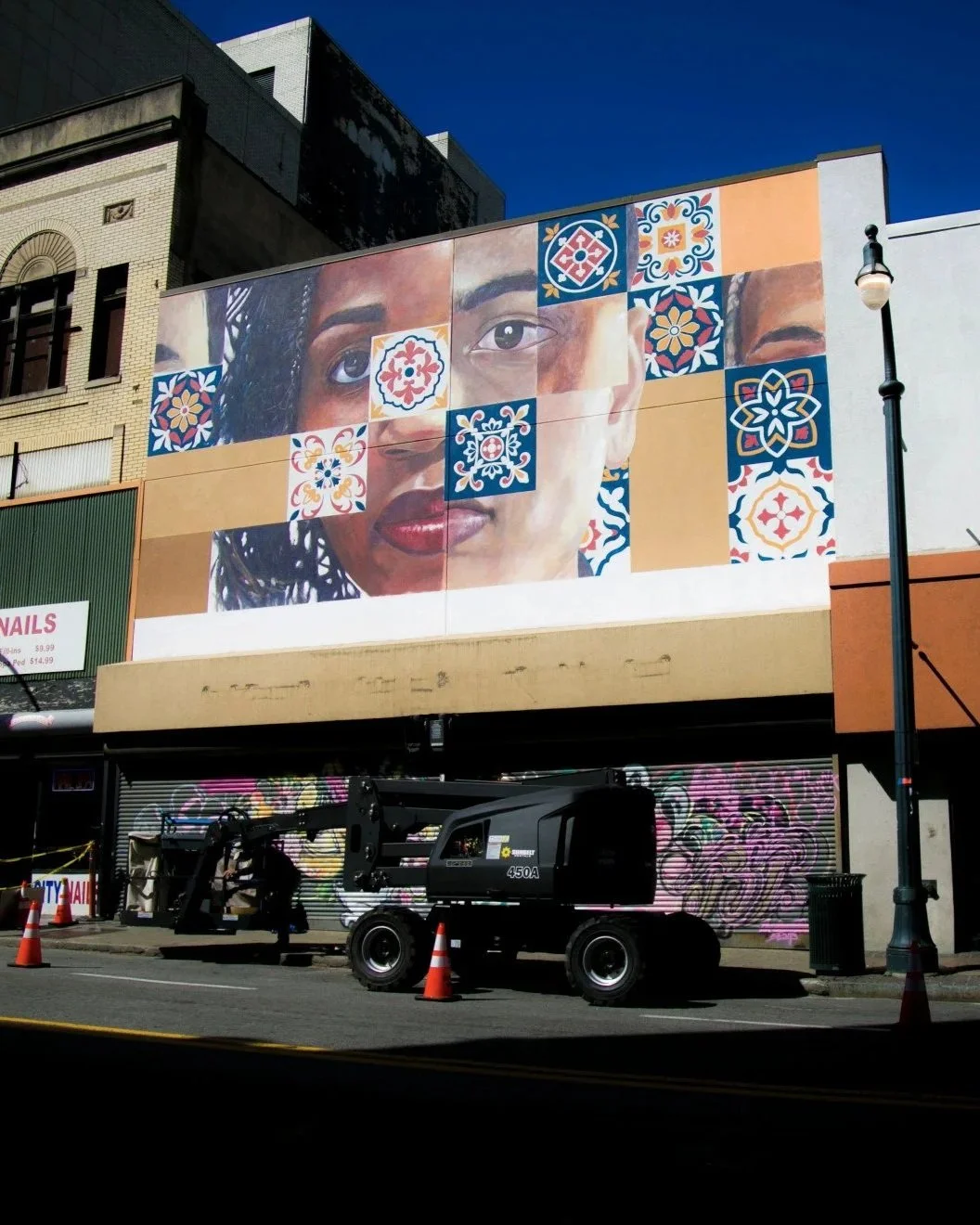 Large mural of a woman’s face with curly hair and dark skin, decorated with colorful patterned tiles around her face, on the side of a building with a dark blue sky in the background.