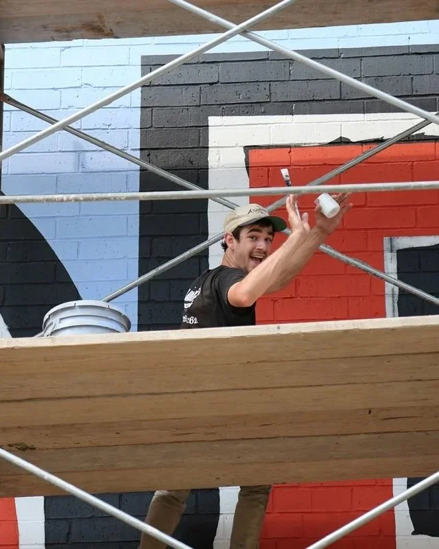 A man painting a mural on a brick wall. He is smiling and holding a paintbrush in one hand and a small container in the other. The wall is painted with geometric shapes in blue, black, white, and red.