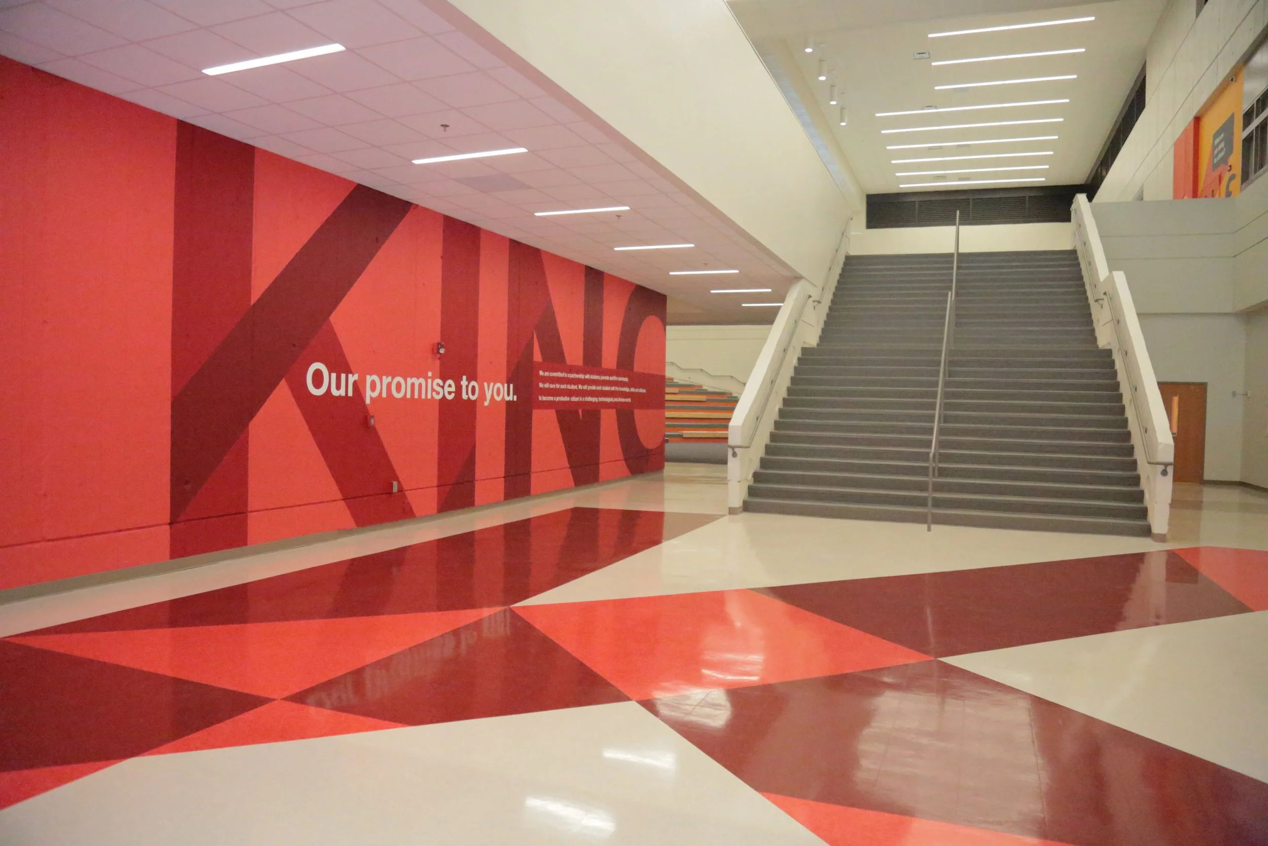 Empty shopping mall with red and beige patterned floor, red wall with large text, and stairs leading to upper level.