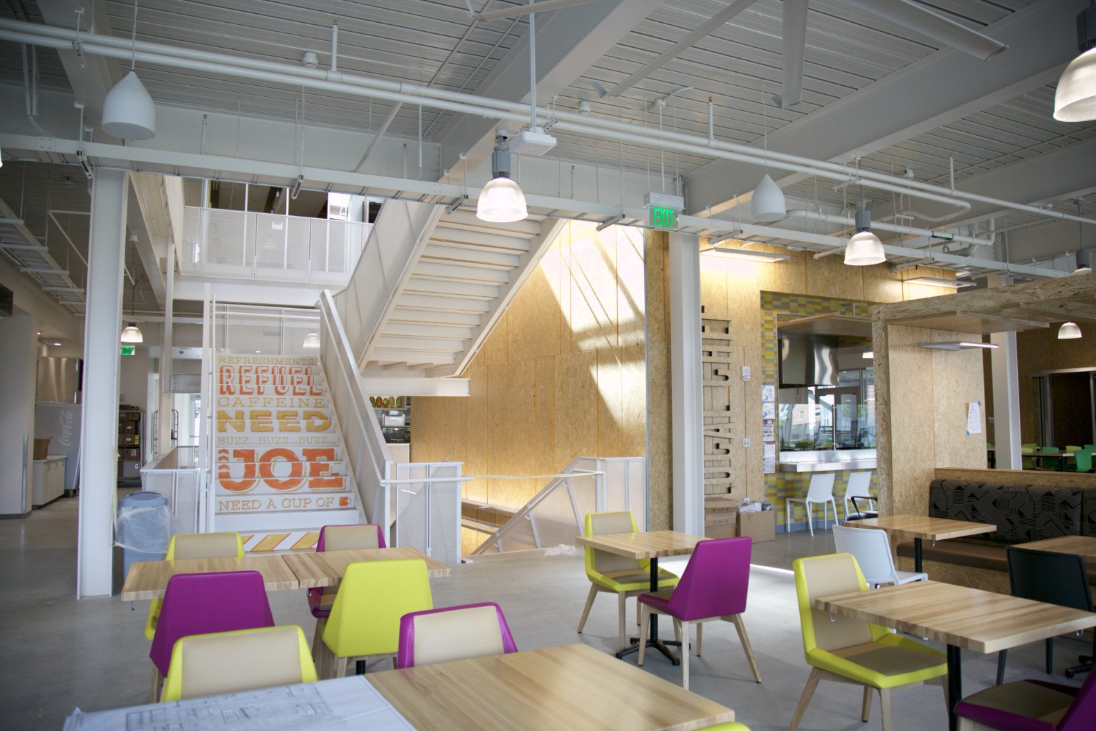Interior of a modern café or restaurant with colorful chairs, wooden tables, a staircase, and natural light coming through large windows.