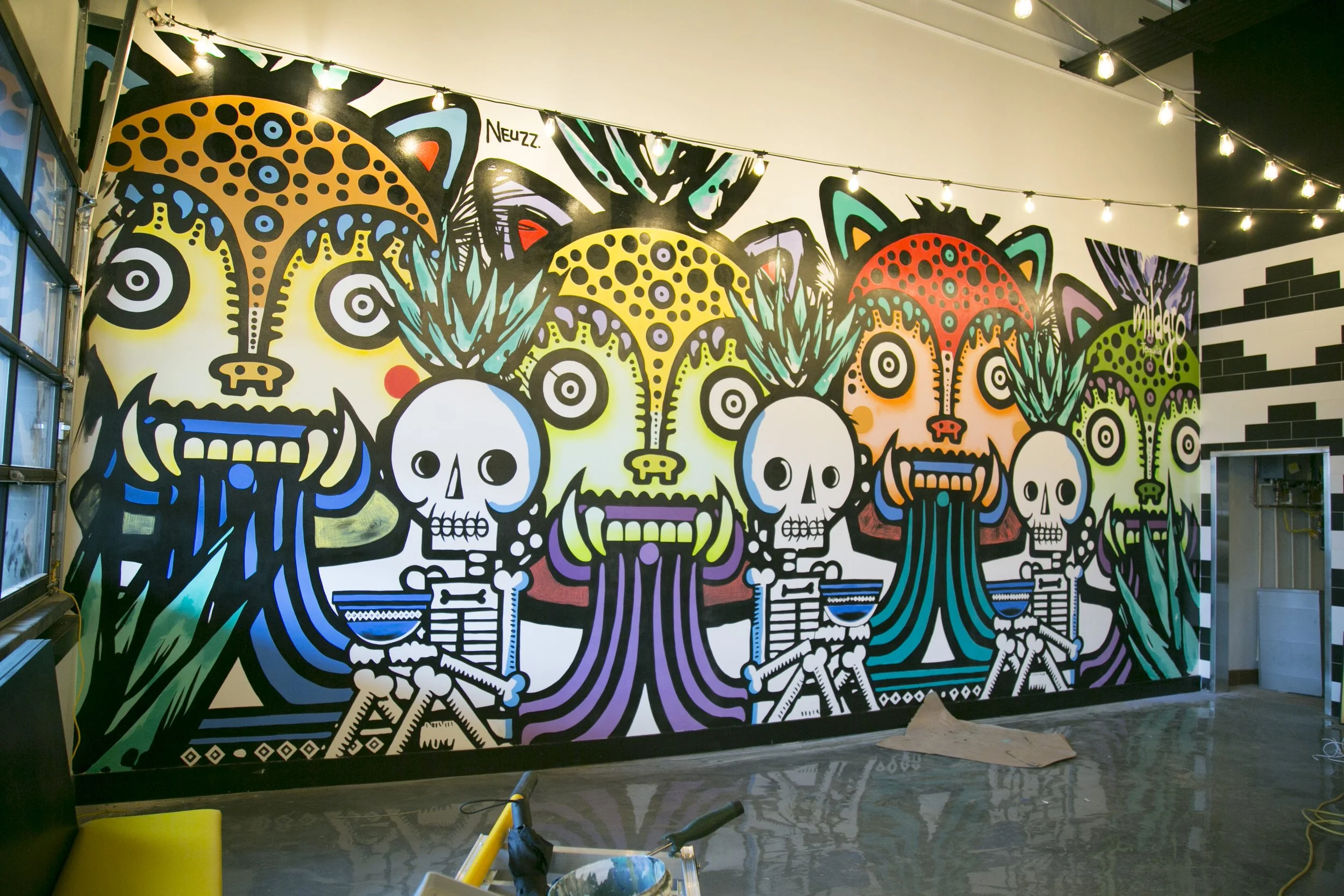 Colorful mural with skulls, animal faces, and abstract patterns on a wall inside a building, with string lights overhead.